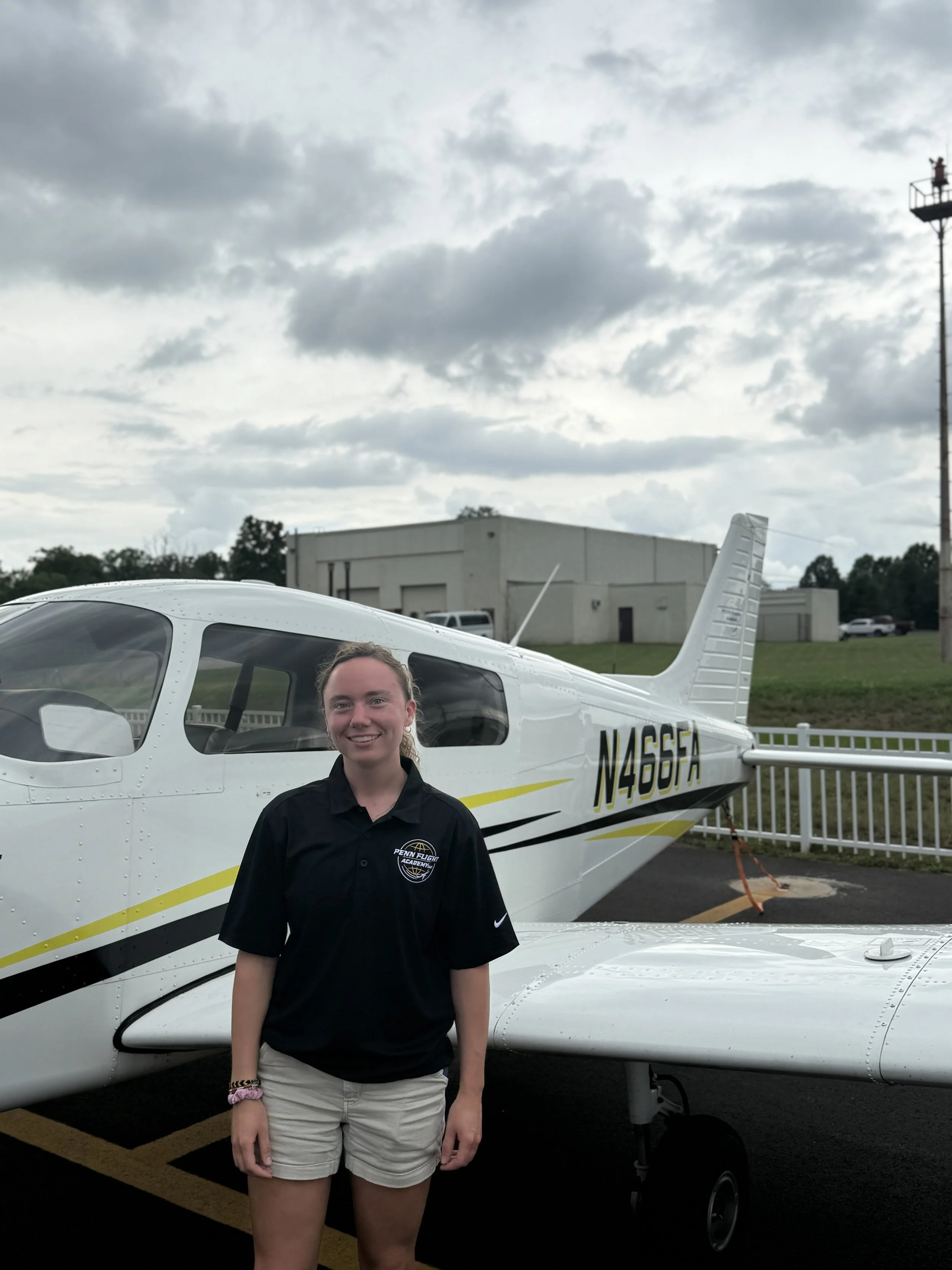 Smiling person wearing a black "Penn Flight School" polo shirt, standing outdoors with sunglasses hanging on the shirt.