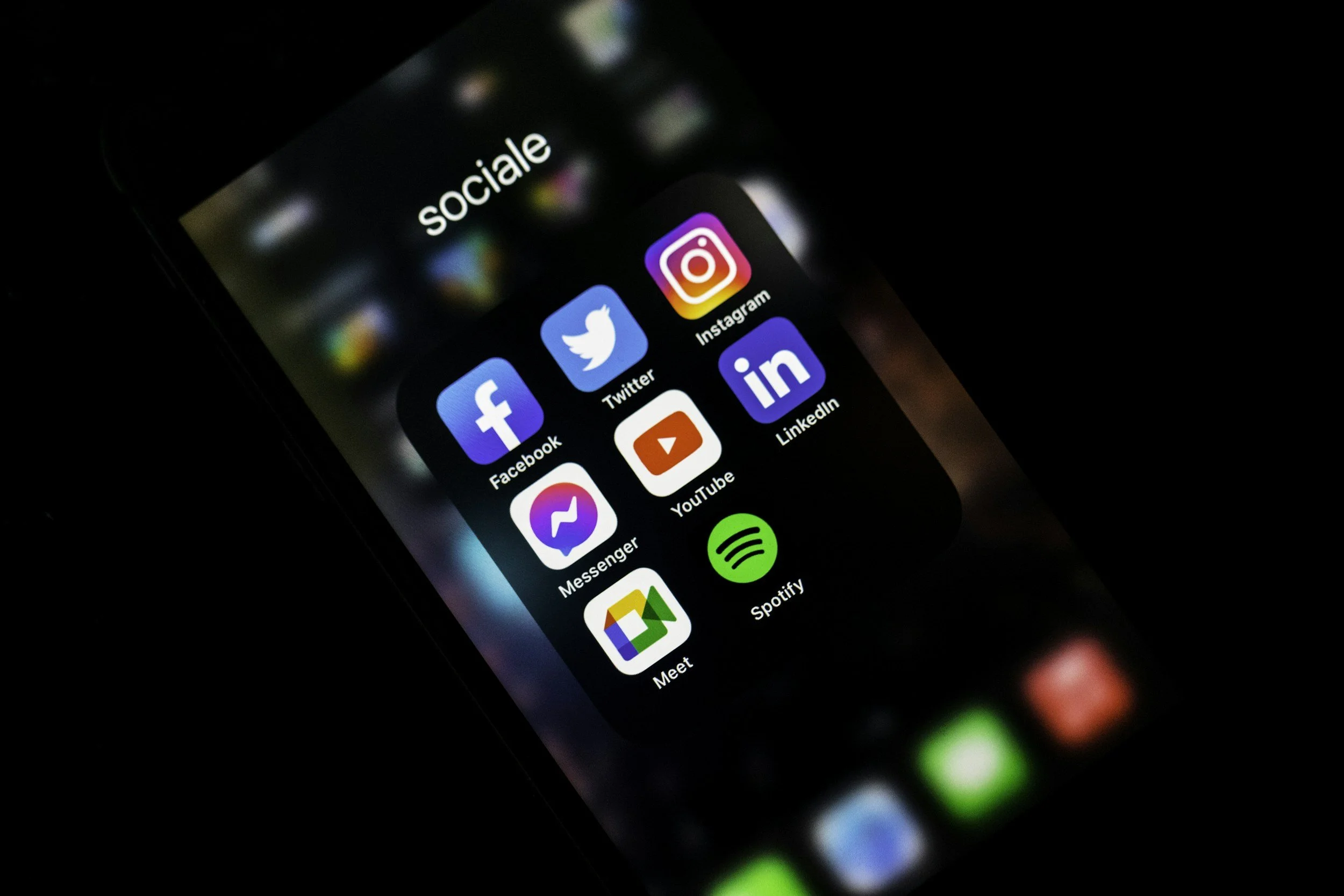 Smartphone screen displaying social media app icons including Facebook, Twitter, Messenger, YouTube, Instagram, LinkedIn, Spotify, and Google Meet, within a folder titled 'sociale' against a dark background.