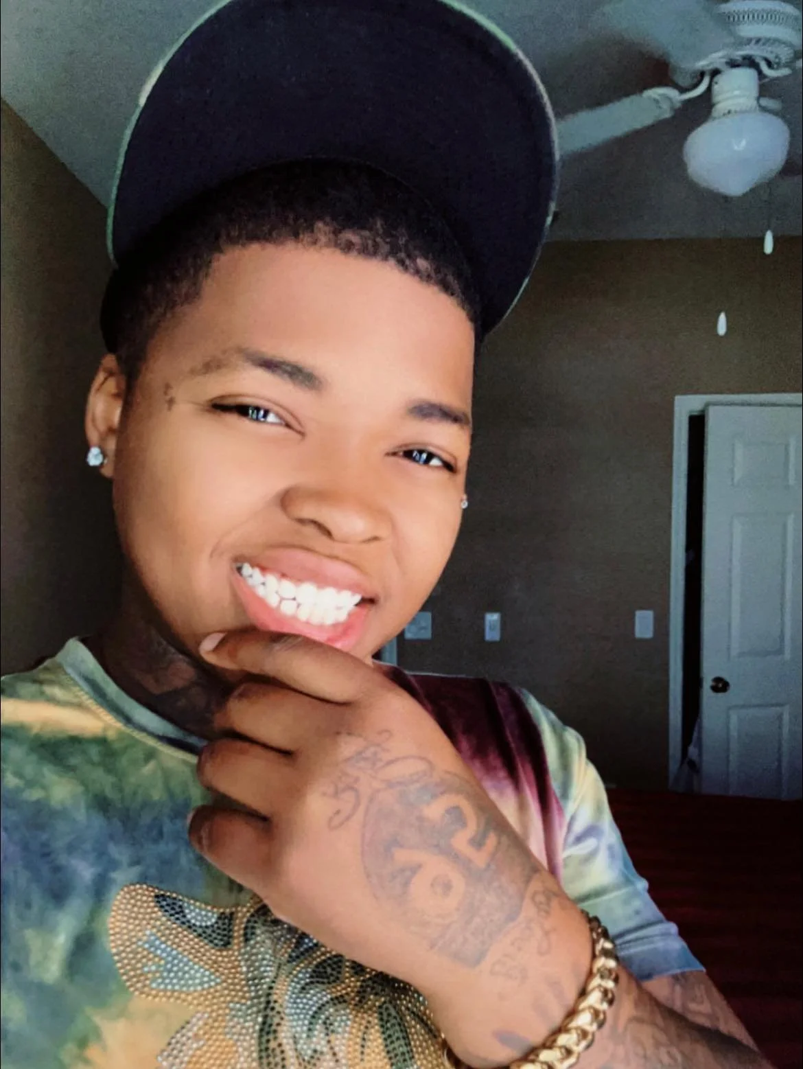 A young person with short curly hair, earrings, and tattoos posing with a smile, wearing a tie-dye shirt and a black cap, in a room with a brown wall and a ceiling fan.