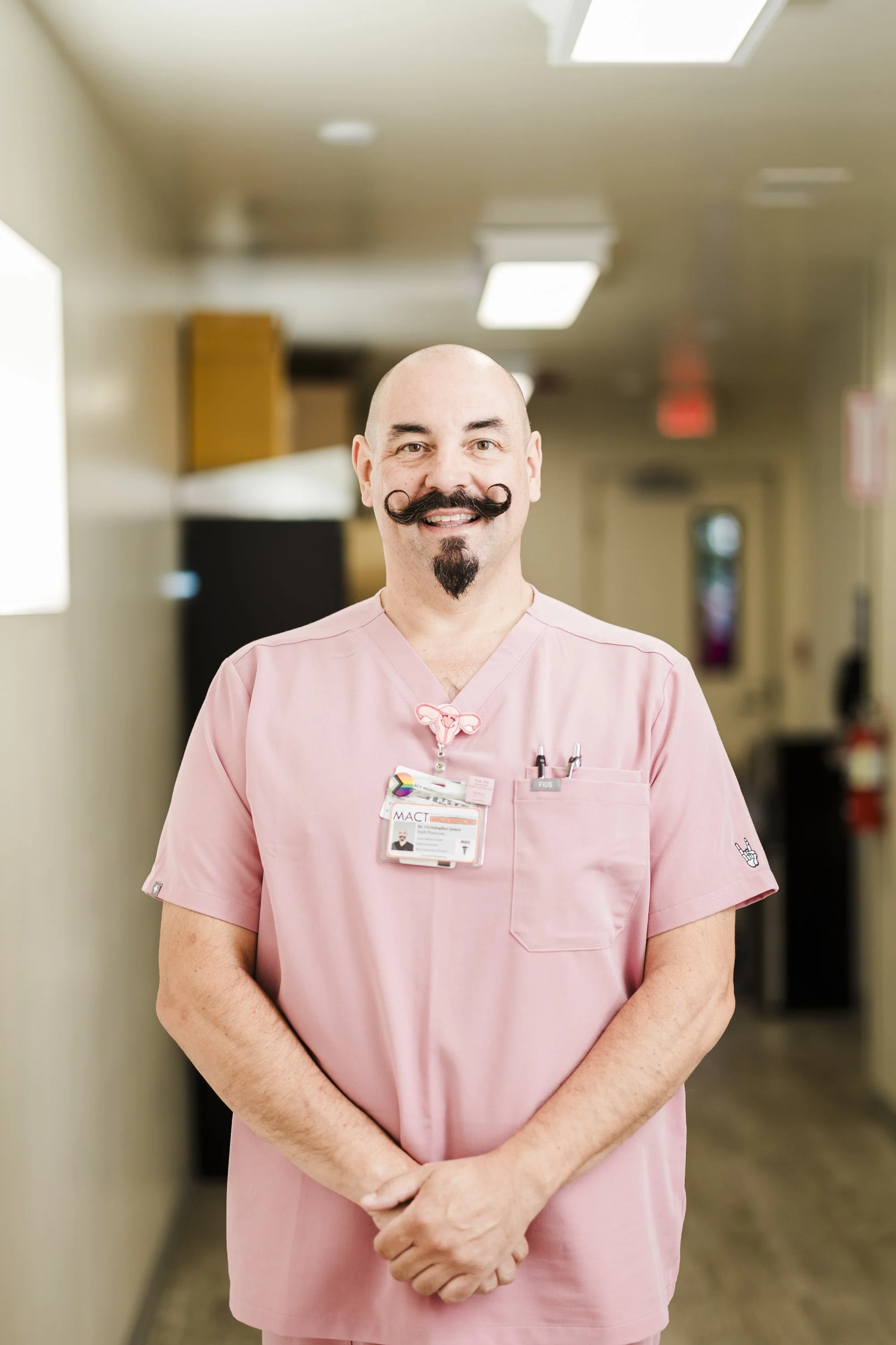 A male nurse standing in a hospital room, wearing pink scrubs and smiling at the camera.