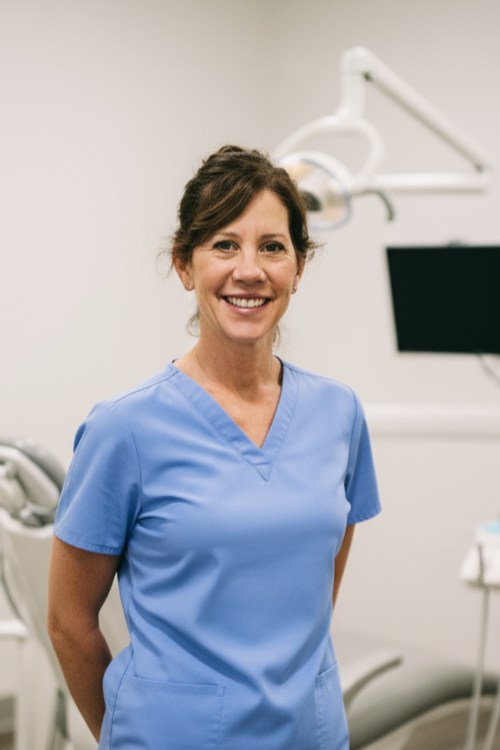 A smiling female healthcare professional in blue scrubs standing in a modern medical office with dental equipment in the background.