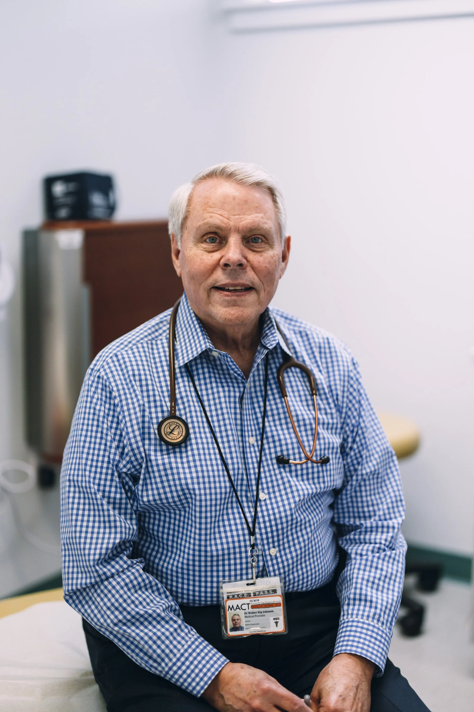 A cheerful male doctor with gray hair, wearing a white coat and a stethoscope around his neck, standing in a medical office or clinic.