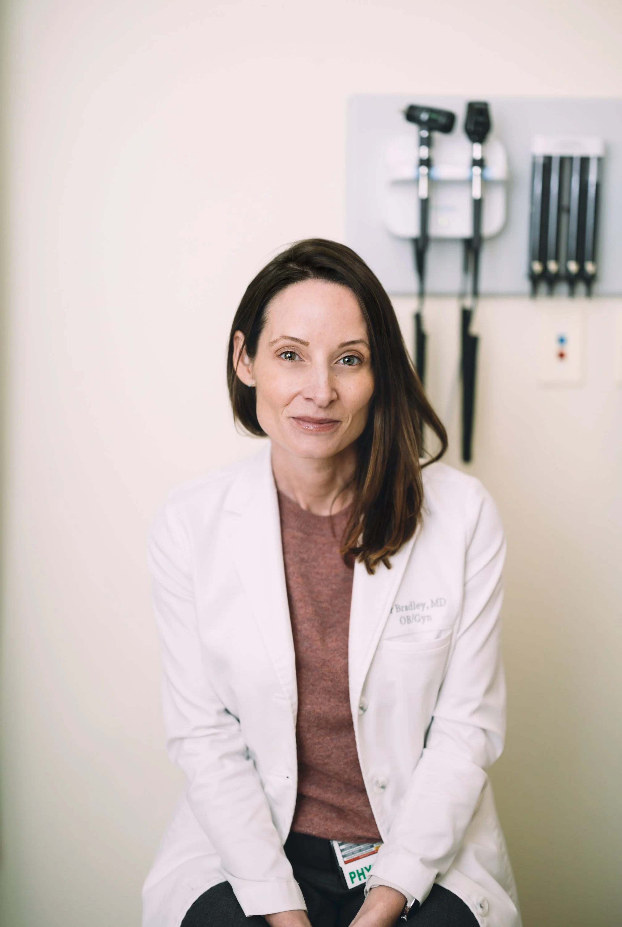 A female doctor wearing a white coat standing in a medical examination room, with medical equipment and an exam table in the background.
