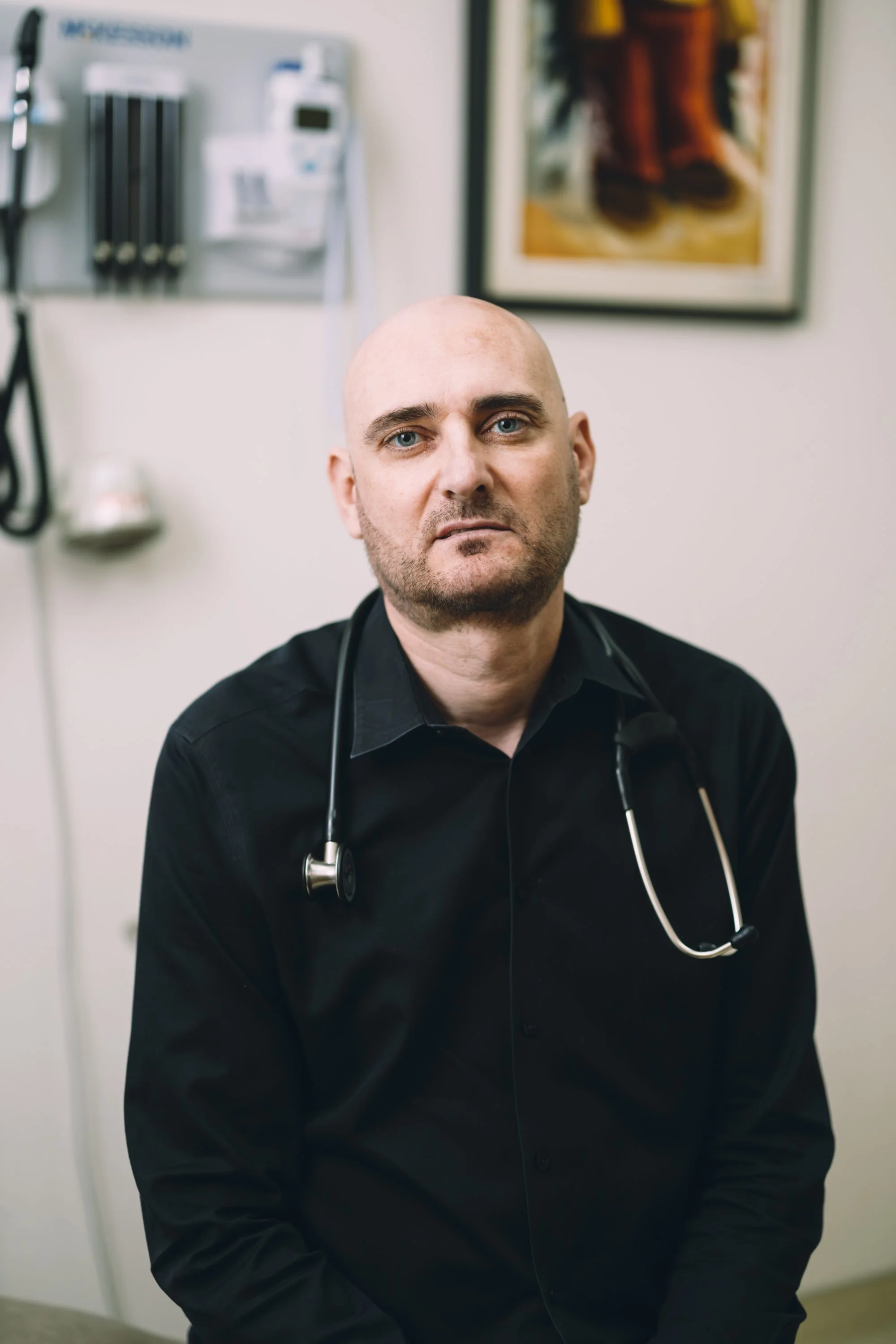 A smiling male doctor with a stethoscope around his neck sitting in a medical office.