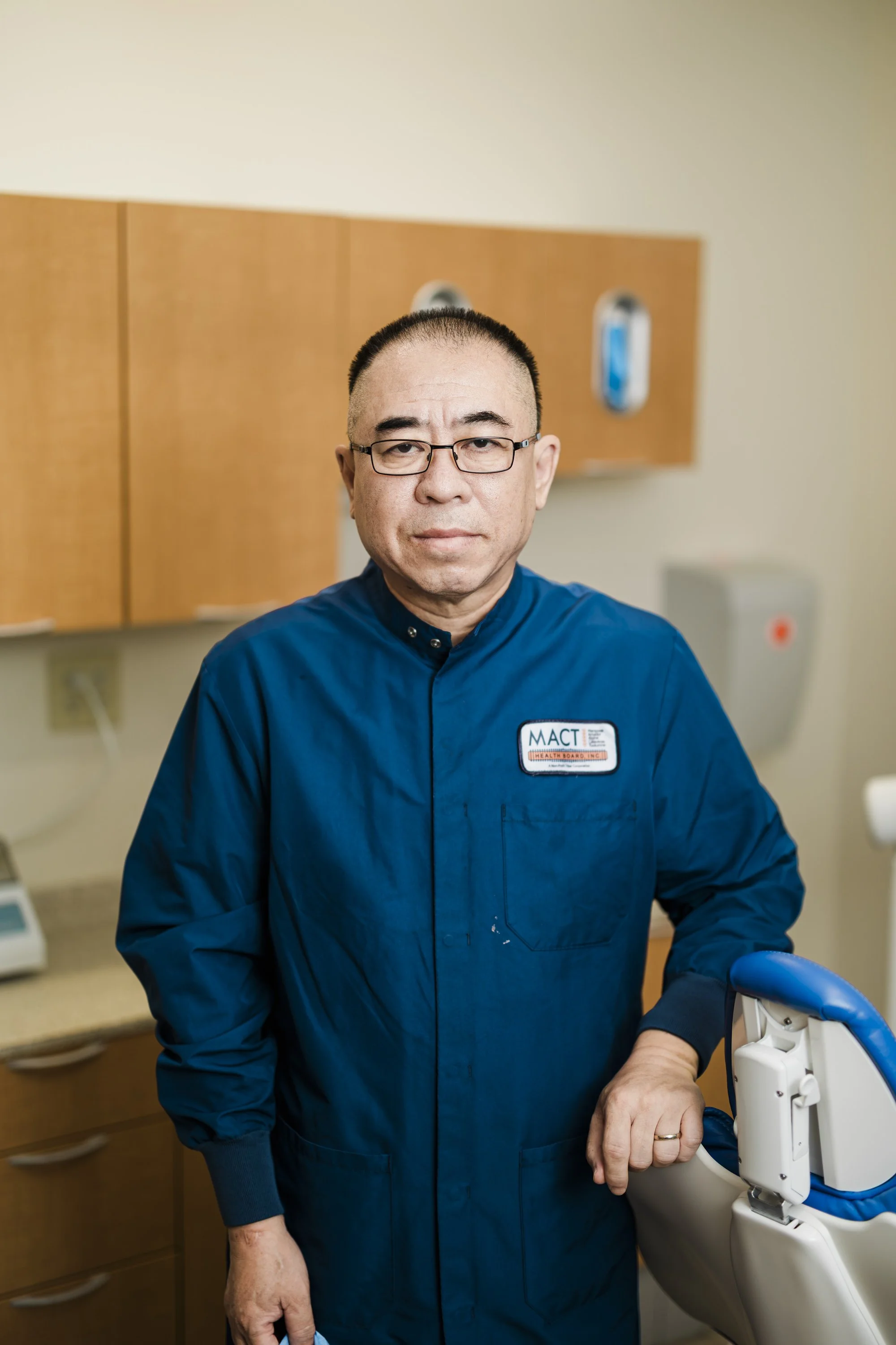 A smiling male healthcare professional wearing glasses and a blue uniform with a badge, standing in a medical office. He is next to a dental chair.