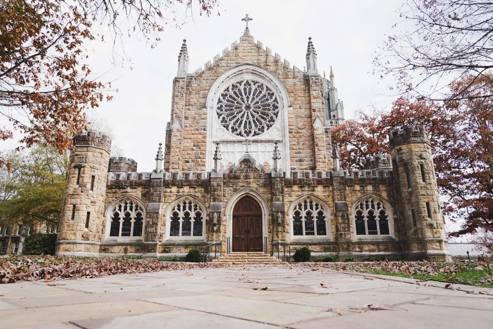 Front view of a historic stone church with a large round stained glass window, decorative turrets, and a wooden door, surrounded by trees with autumn foliage.
