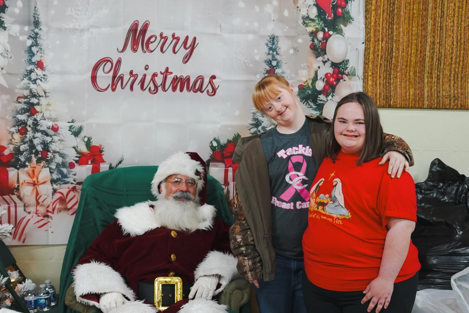 A person dressed as Santa Claus sitting on a green chair with a festive background, along with two young girls smiling and standing next to him, during a Christmas celebration.