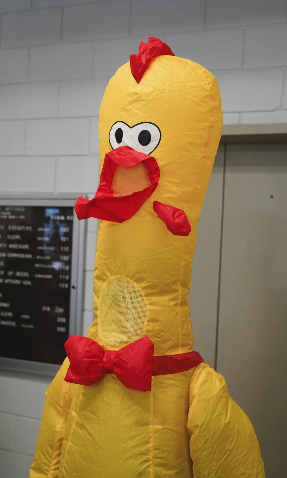 A person dressed in a chicken costume made of yellow paper and red paper accents, including a comb, bow tie, and beak, with cartoonish eyes on the costume's head.