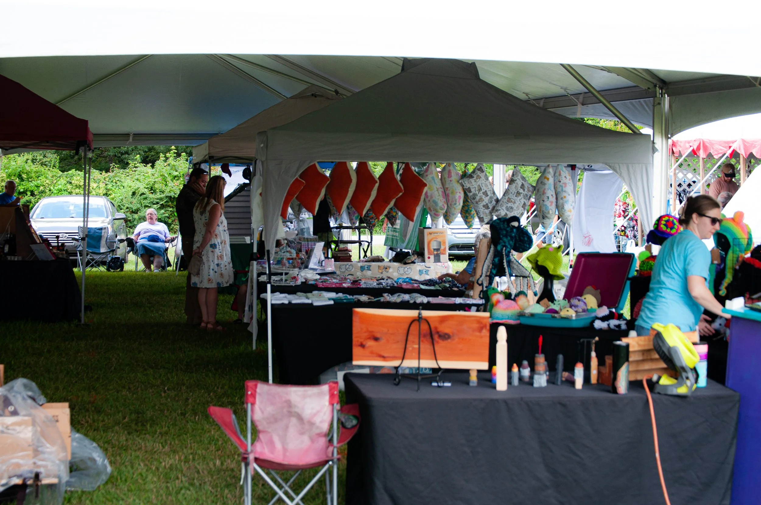 A vendor booth at an outdoor market selling colorful handmade pillows and other crafts. Several people are seen browsing and shopping.