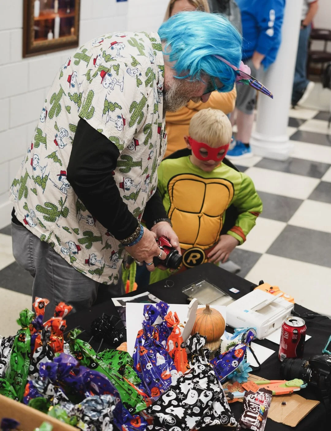 Man with blue hair and colorful shirt looks at Halloween treats on table with child dressed as Teenage Mutant Ninja Turtle, both wearing masks. The table has Halloween-themed items and a small pumpkin.