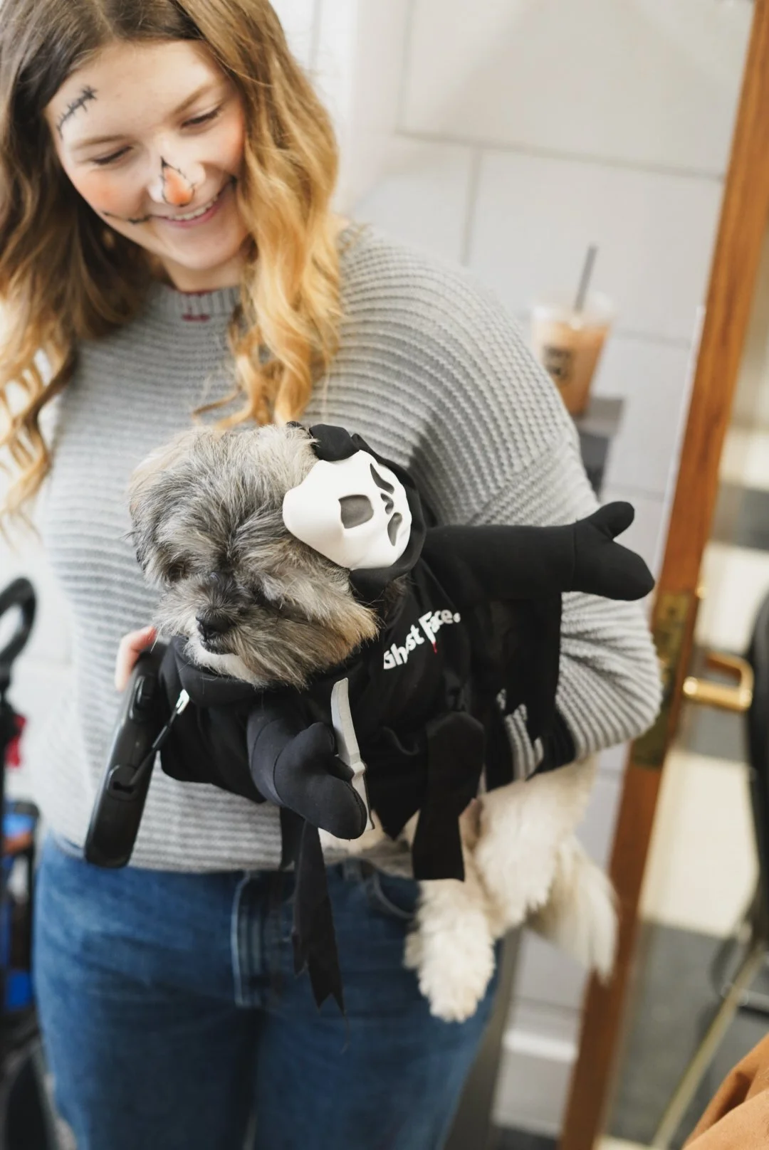 A woman with face paint resembling a skeleton holds a small dog dressed in a ghost face mask and black costume.