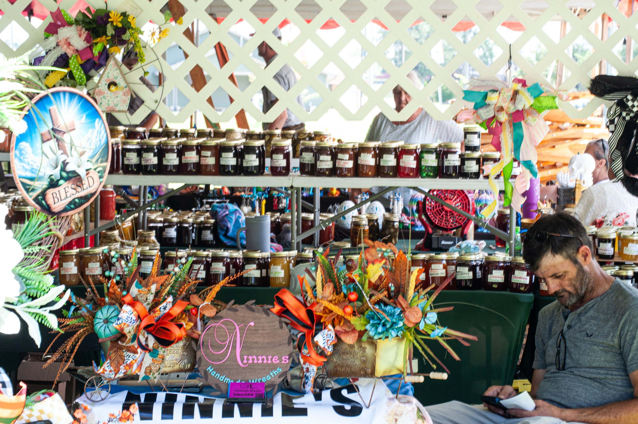 A display at an outdoor craft fair with jars of jam on tables, colorful decorative items including a floral wreath, and people in the background browsing the booths.