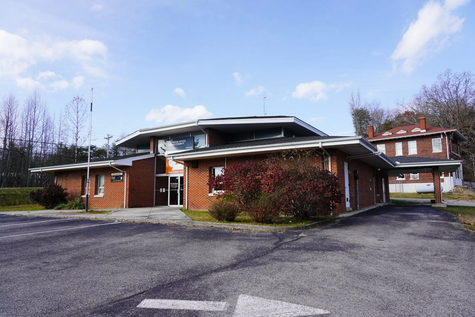 Brick government office building with parking lot and small landscaping, surrounded by trees and a partly cloudy sky.