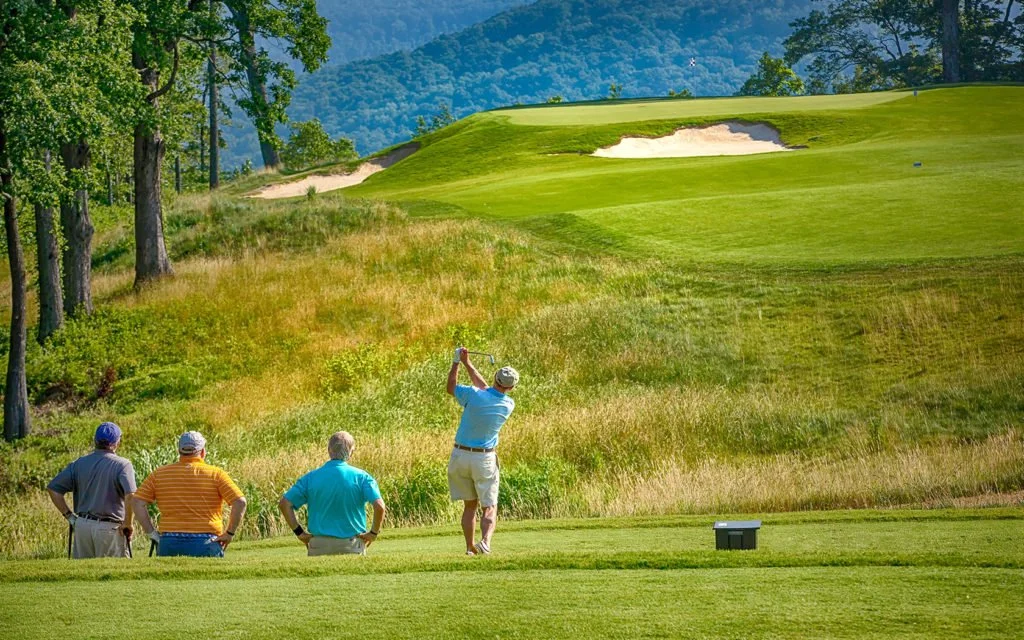 A golfer swings a club on a golf course with three spectators watching. The course has lush green grass, trees, and a sand bunker in the background.