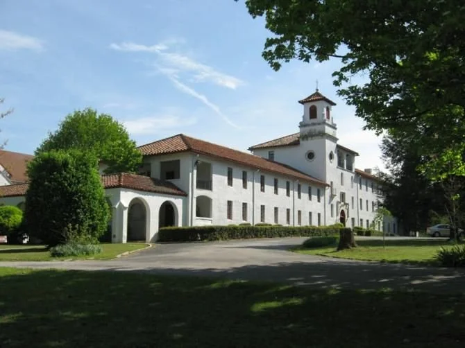 A large white building with a red-tiled roof, surrounded by trees and greenery, under a blue sky with some clouds.