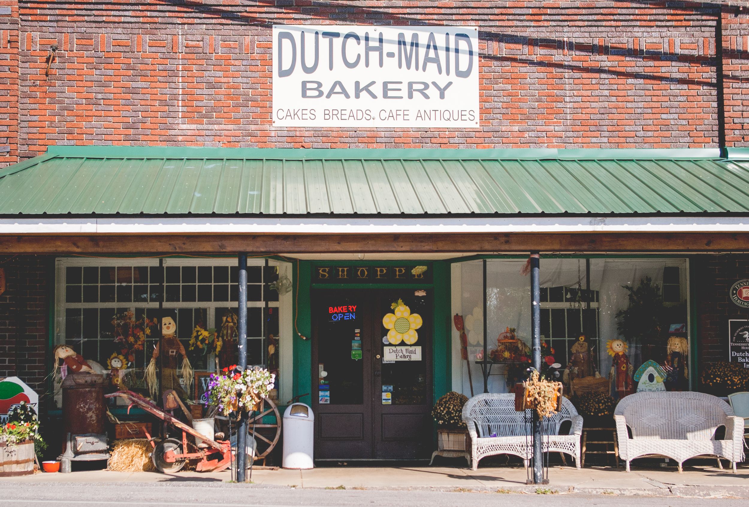 Exterior view of Dutch Maid Bakery with a brick facade, green metal roof awning, decorated with scarecrows, flowers, and wicker furniture, with a sign that reads 'Cakes Breads Cafe Antiques'.