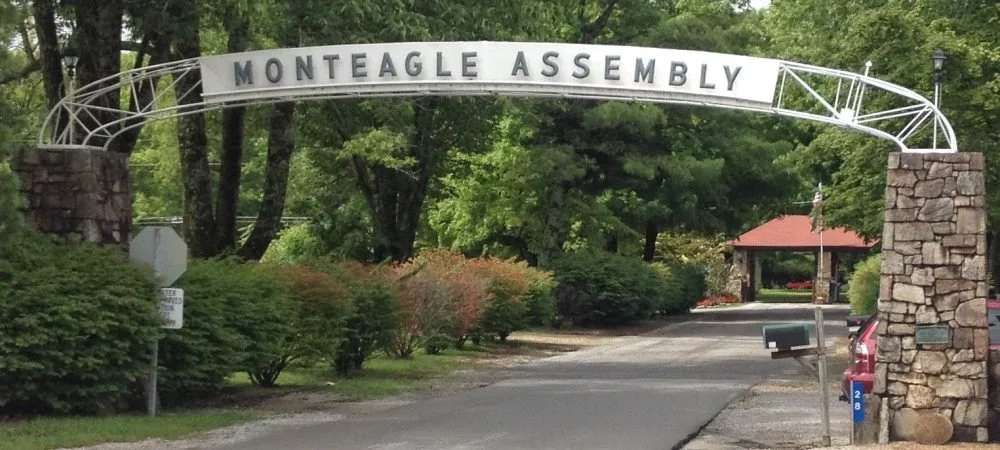 Entrance sign for Monteagle Assembly, with a white arched sign on metal framework supported by stone columns, green trees and shrubs in the background.