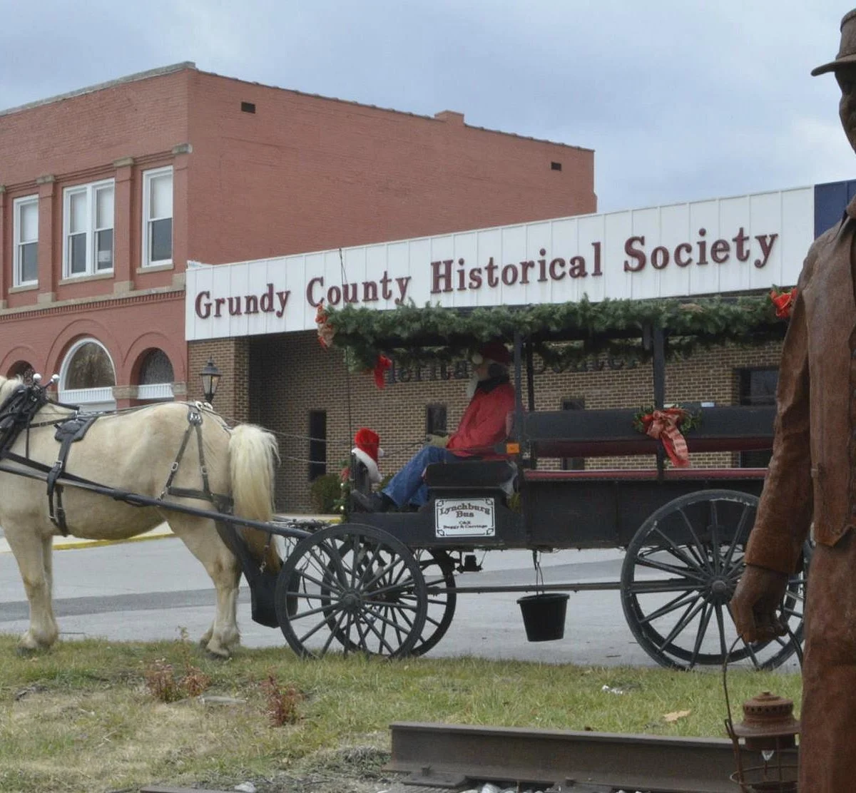 Horse-drawn carriage decorated with holiday greenery parked in front of a brick building with a sign that reads 'Gundy County Historical Society'.