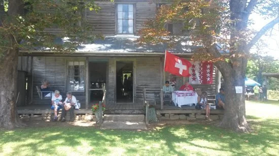 People sitting on a porch of a historic wooden house decorated with Swiss flags, with trees and grass in front of it.