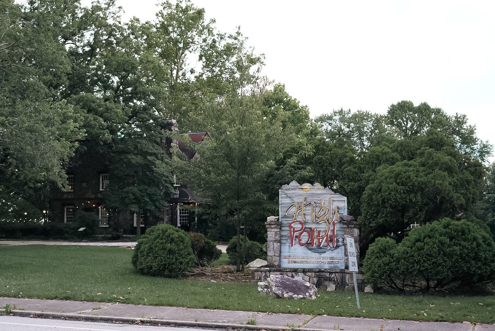 A stone sign in front of a wooded area reads 'High Point' with additional text about historic dining locations on the summit between Chicago and Miami. Behind the sign are large trees and a dark, old-style building with a red roof, partially obscured