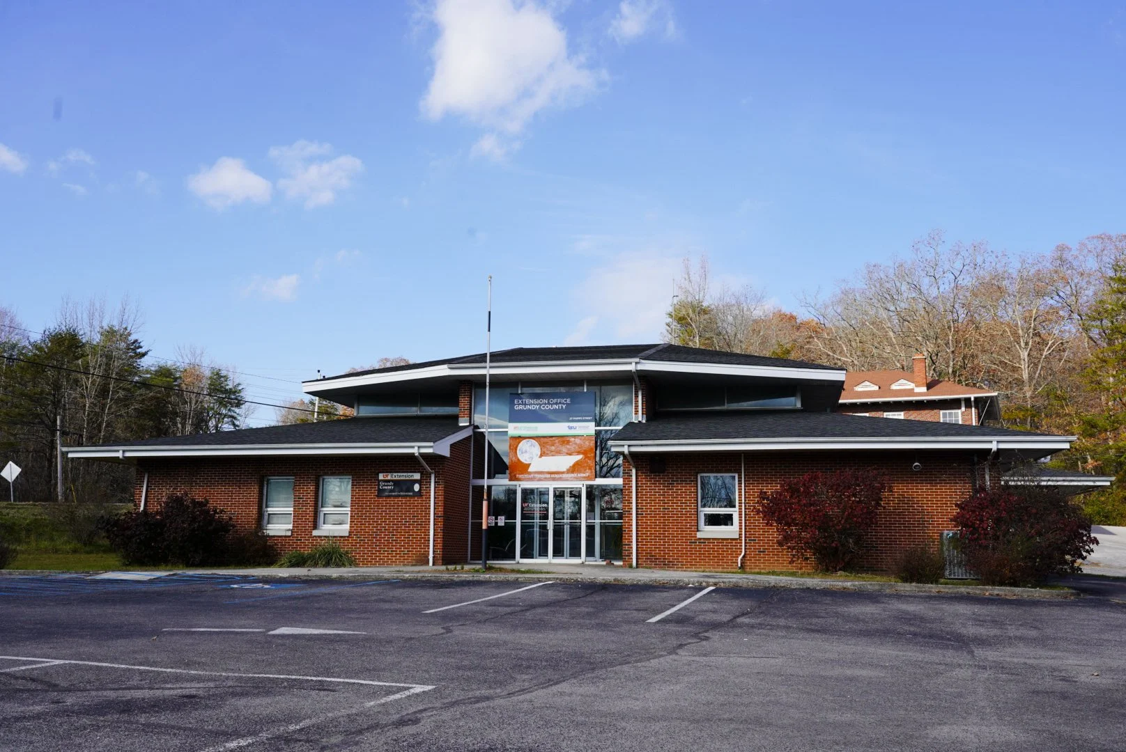 A two-story brick building with a sign that reads 'Extension Office Groundy County' and a parking lot in front, surrounded by trees with fall foliage in the background, under a partly cloudy sky.