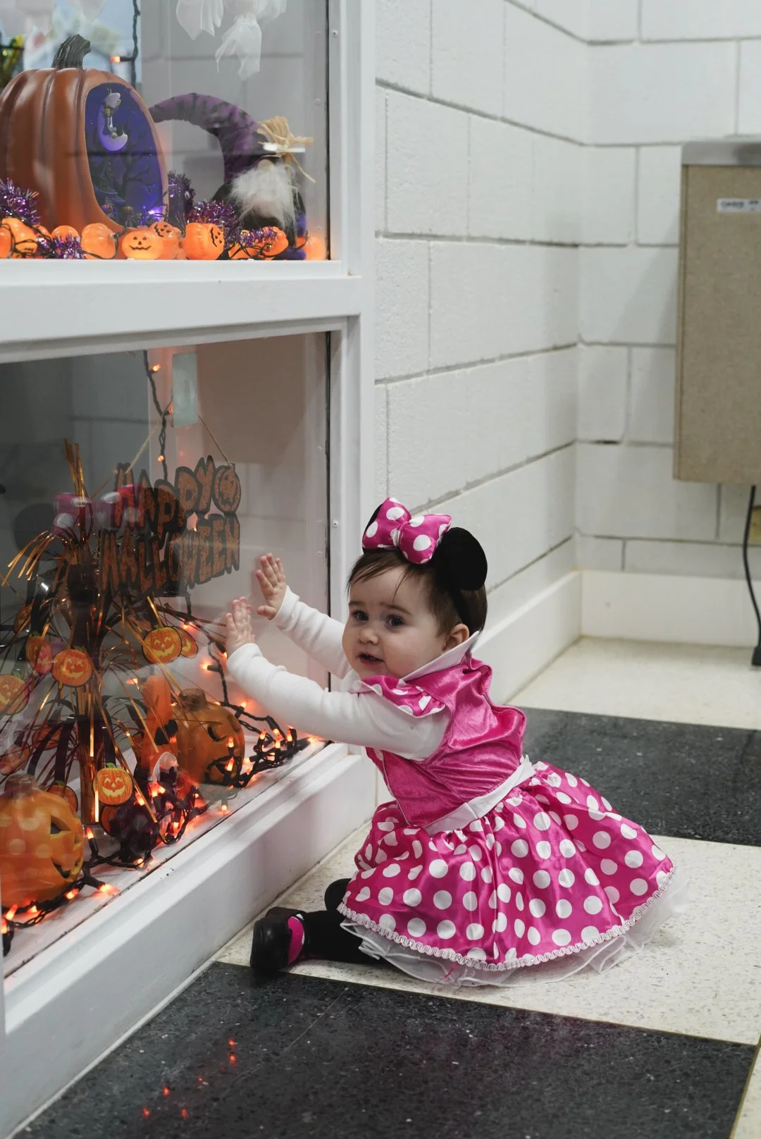 Baby girl dressed as Minnie Mouse touching a Halloween-themed window display with pumpkins and decorations.