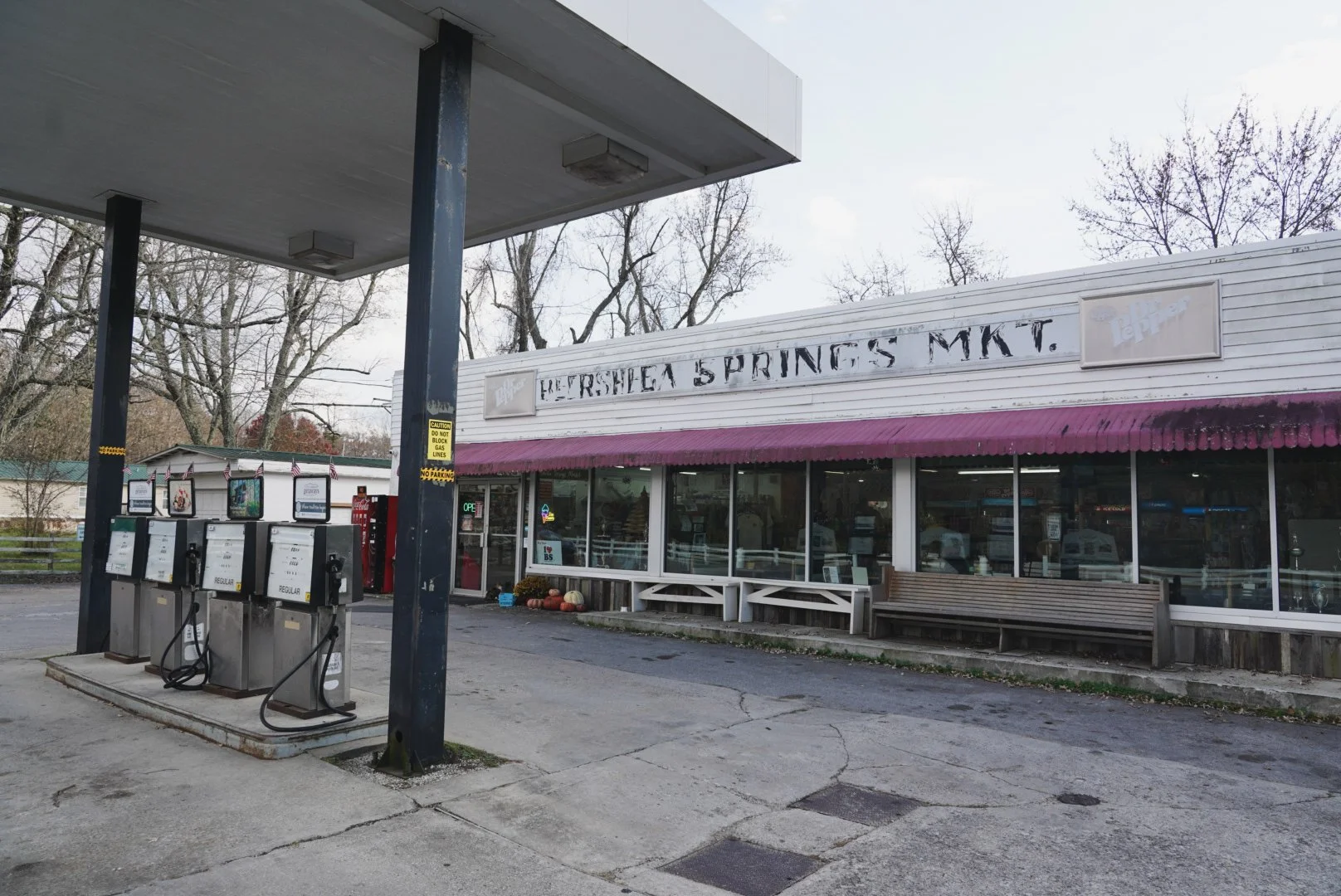 Gas station in front of a store named 'Refresha Springs Mkt.' with pumpkins outside, gas pumps in the foreground, and trees with bare branches in the background.