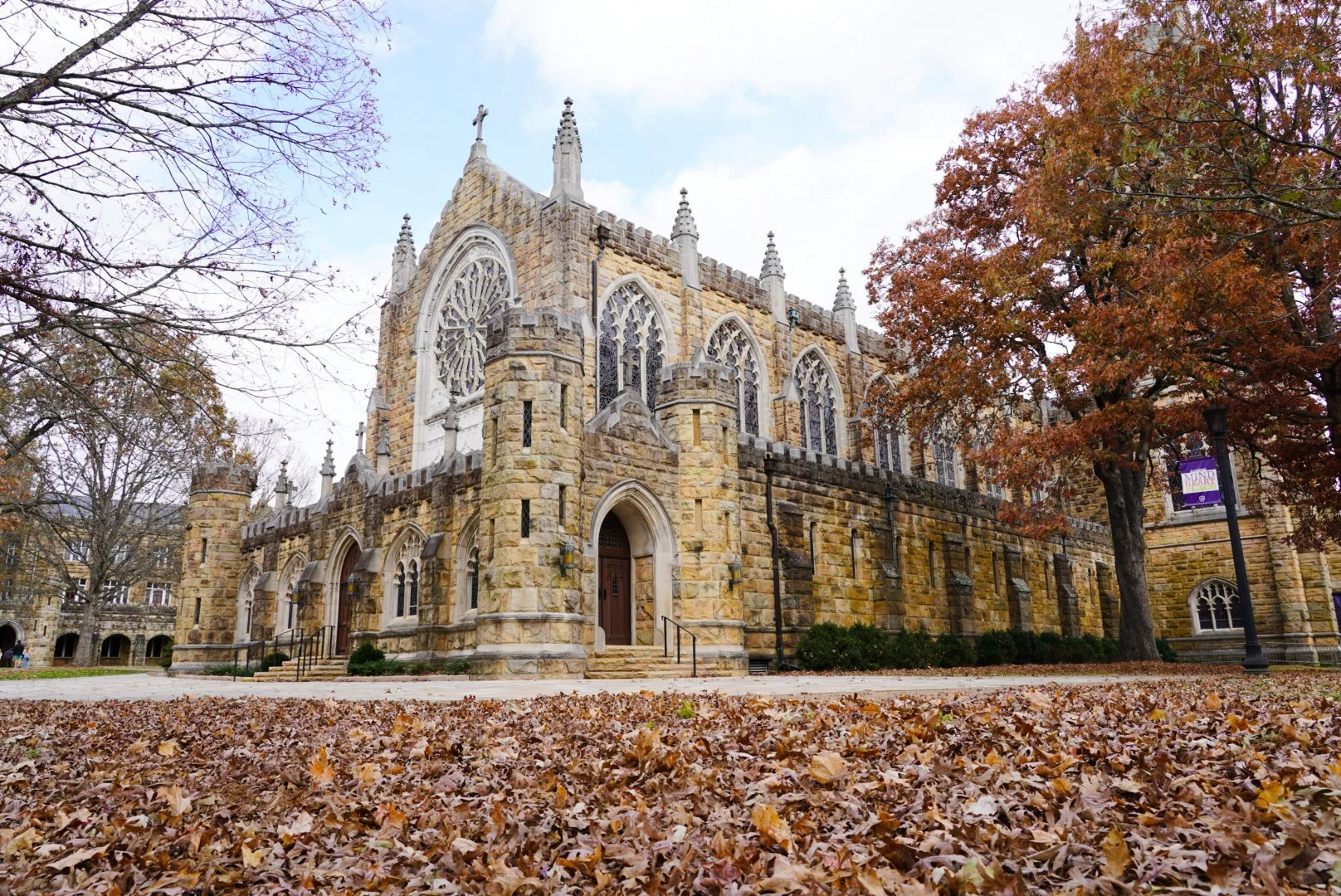 A historic stone church with Gothic architecture, pointed arches, and large stained glass windows, surrounded by autumn trees and fallen leaves.