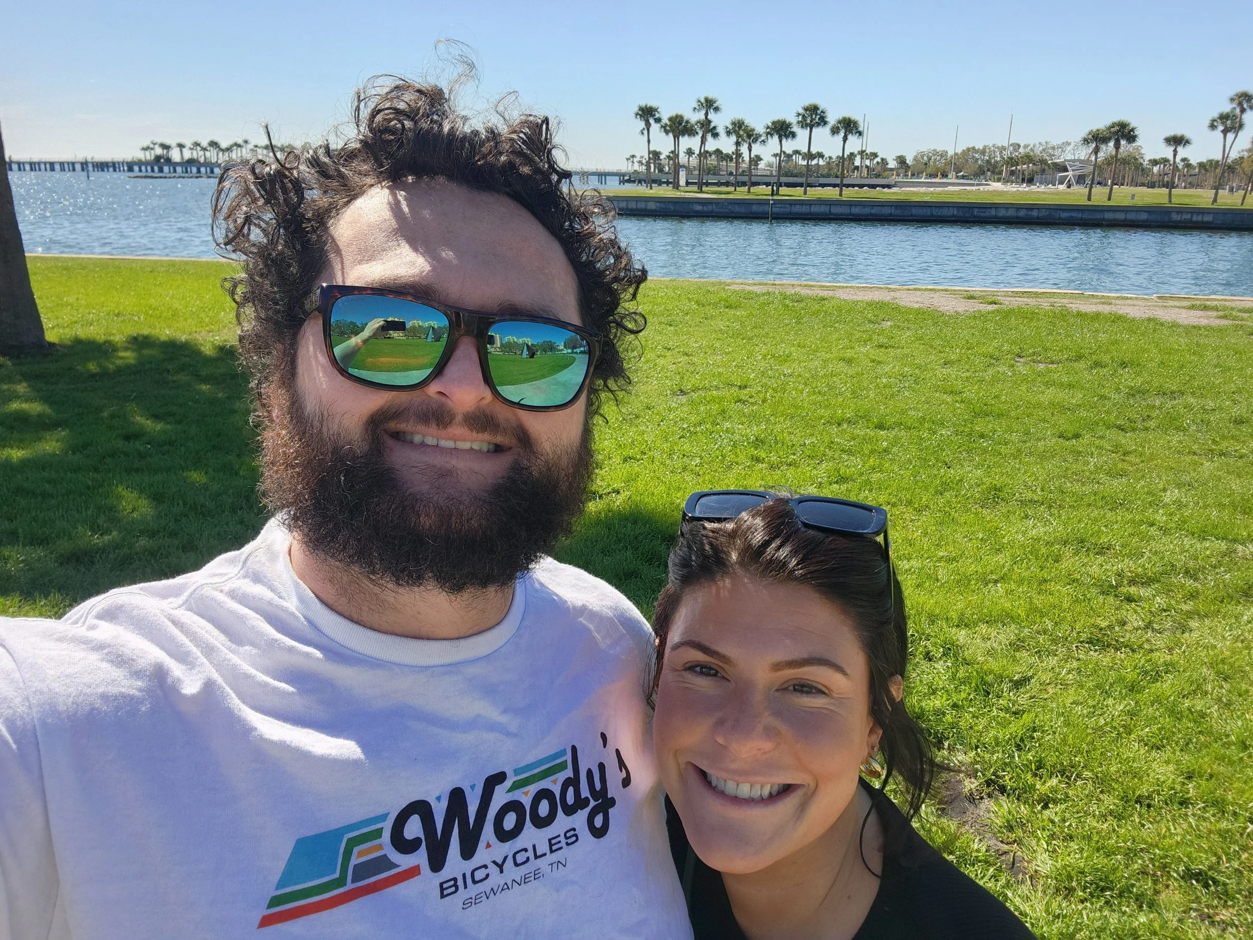 A smiling man with curly hair and a beard wearing sunglasses and a white t-shirt with Woody's Bicycles logo, and a smiling woman with dark hair and sunglasses on her head, pose for a selfie outdoors near a waterway with palm trees and a clear blue sky in the background.