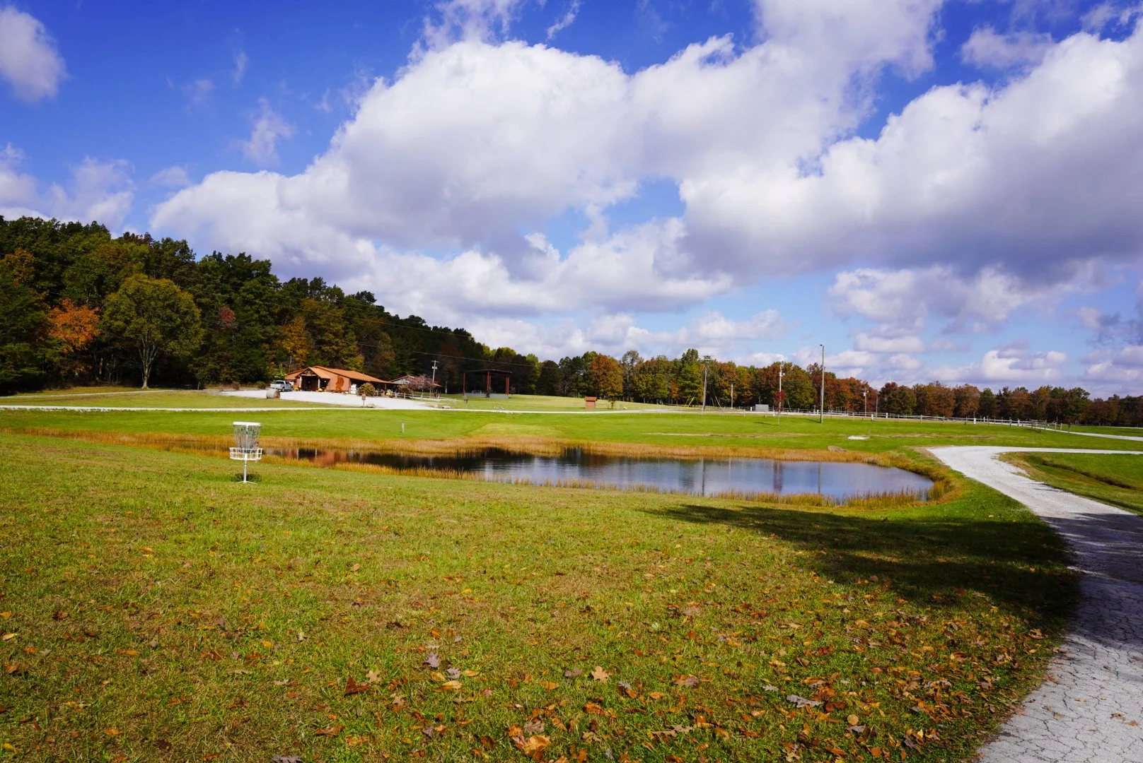 A park with a small pond, disc golf basket, grassy area, and a dirt walking path. In the background, there is a building surrounded by trees with autumn foliage. The sky is partly cloudy with white fluffy clouds and some blue patches.