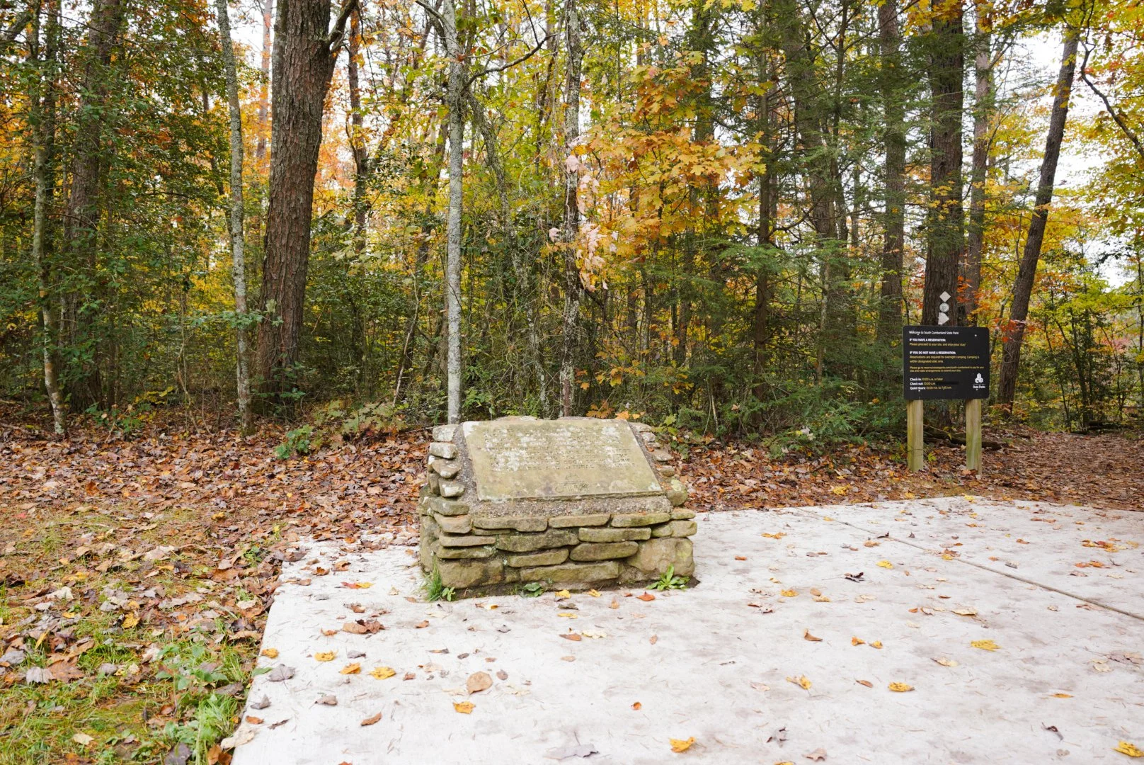 A historical marker and a stone memorial in a wooded area with fallen leaves on the ground and orange, yellow, and green foliage on the trees.