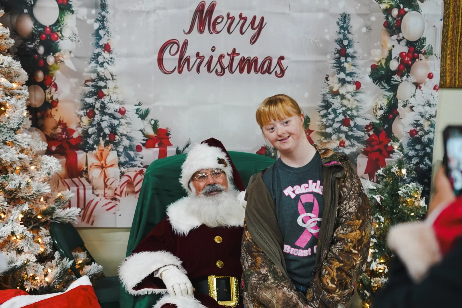 A young girl with red hair and a camouflage jacket posing with Santa Claus during Christmas celebrations. Santa is seated on a green chair, wearing a red suit with white fur trim, glasses, and a Santa hat. The background features Christmas trees deco