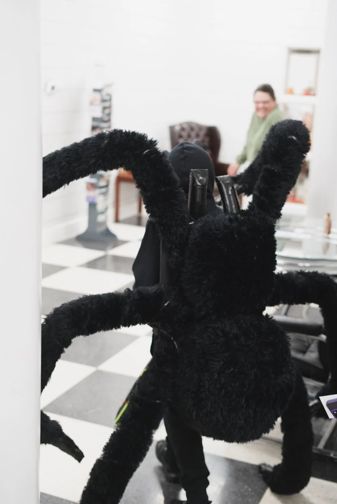 A fuzzy black spider-shaped stuffed toy with a shiny black plastic face and several plastic tubes on its back, placed on a desk in an office setting. In the background, a woman with glasses and a green shirt is smiling, sitting in a brown armchair.