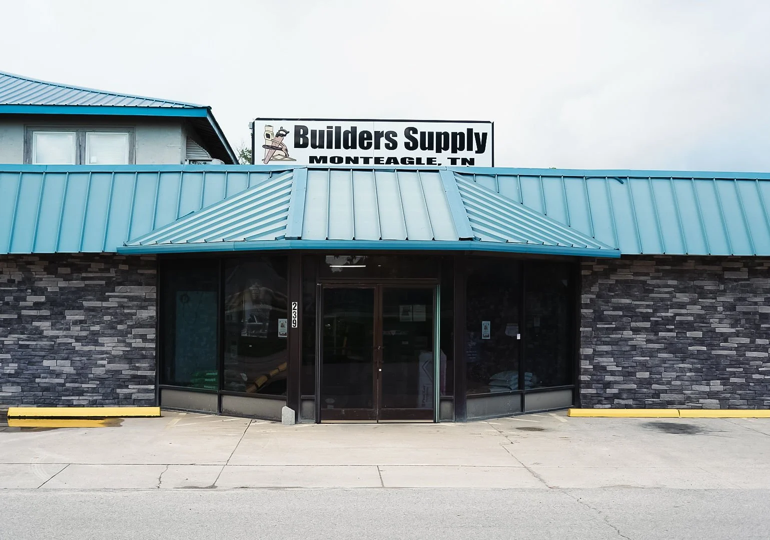 Front view of a building with a stone facade and a blue metal roof, with a sign on top that reads 'Builders Supply, Monteagle, TN.' The entrance has large glass doors and windows, with a sidewalk and yellow parking lot barriers in front.