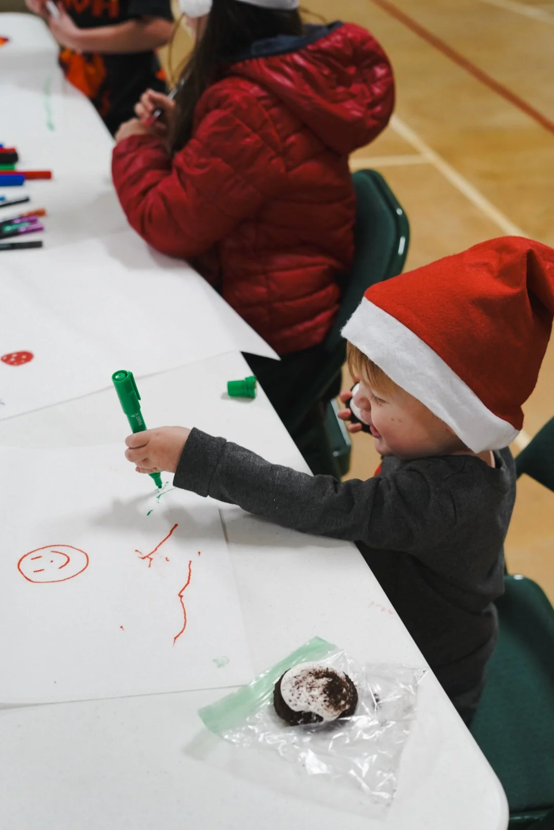 A young boy wearing a Santa hat is drawing with a green marker on a large sheet of white paper, smiling as he colors. Other children are sitting at the table, which is decorated with markers, and a candy treat wrapped in clear plastic is on the table