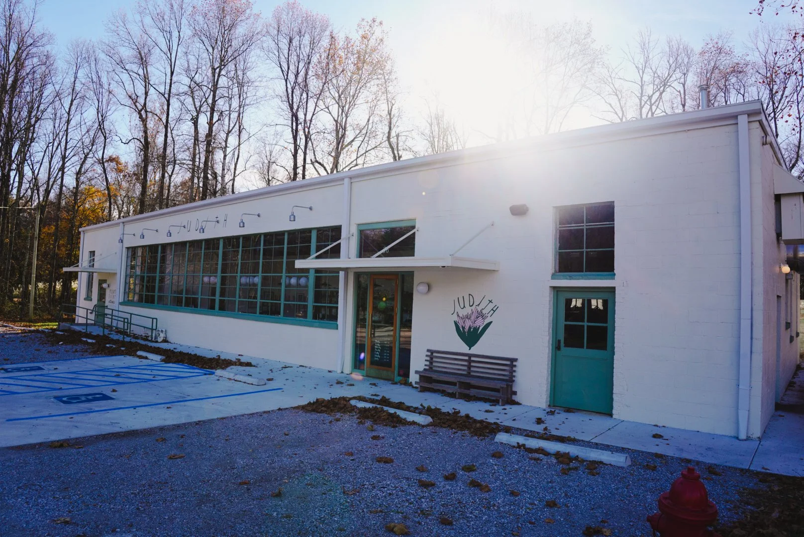 Exterior of a white building with a large glass window, teal door, and a painted flower with the words "JUDITH" on the wall. There is a black bench, a parking lot with blue Handicap markings, and trees in the background.