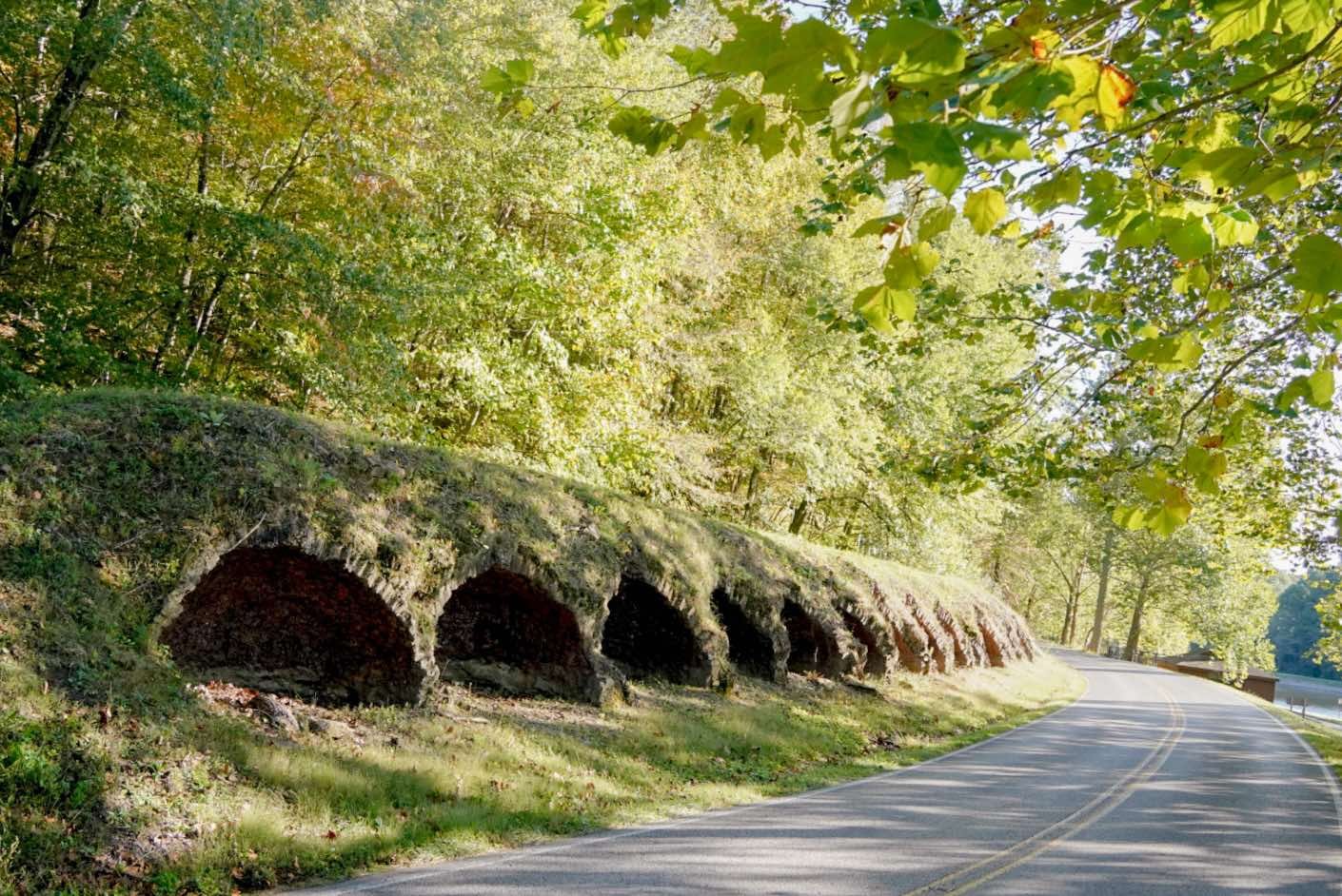 A scenic road winding through a forest with green leaves, featuring a historic stone bridge with multiple arches on the side of the road.