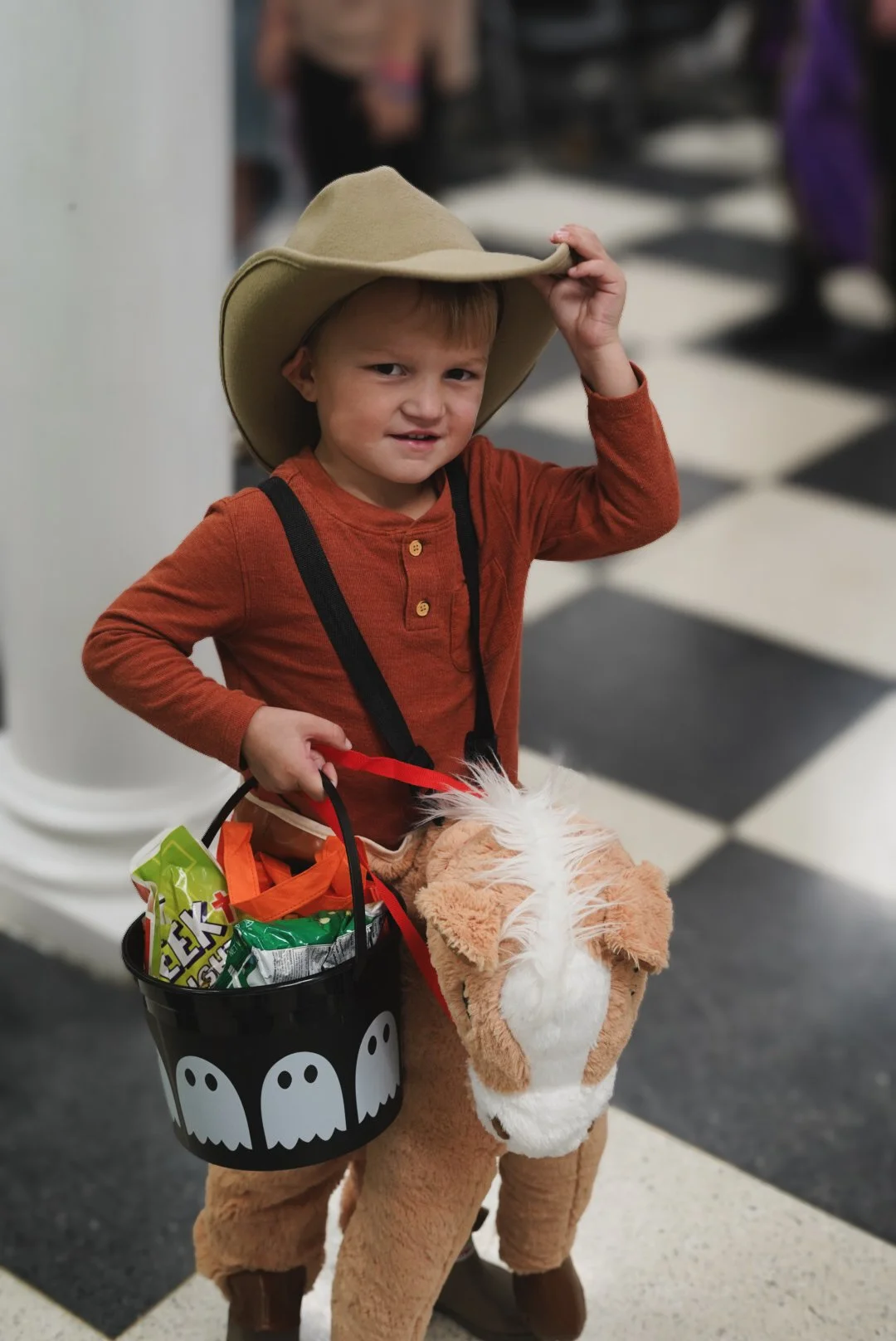 Young boy dressed in Halloween costume, wearing a cowboy hat and riding a plush horse toy, holding a Halloween-themed trick-or-treat bag filled with snacks, inside a building with black and white checkered flooring.