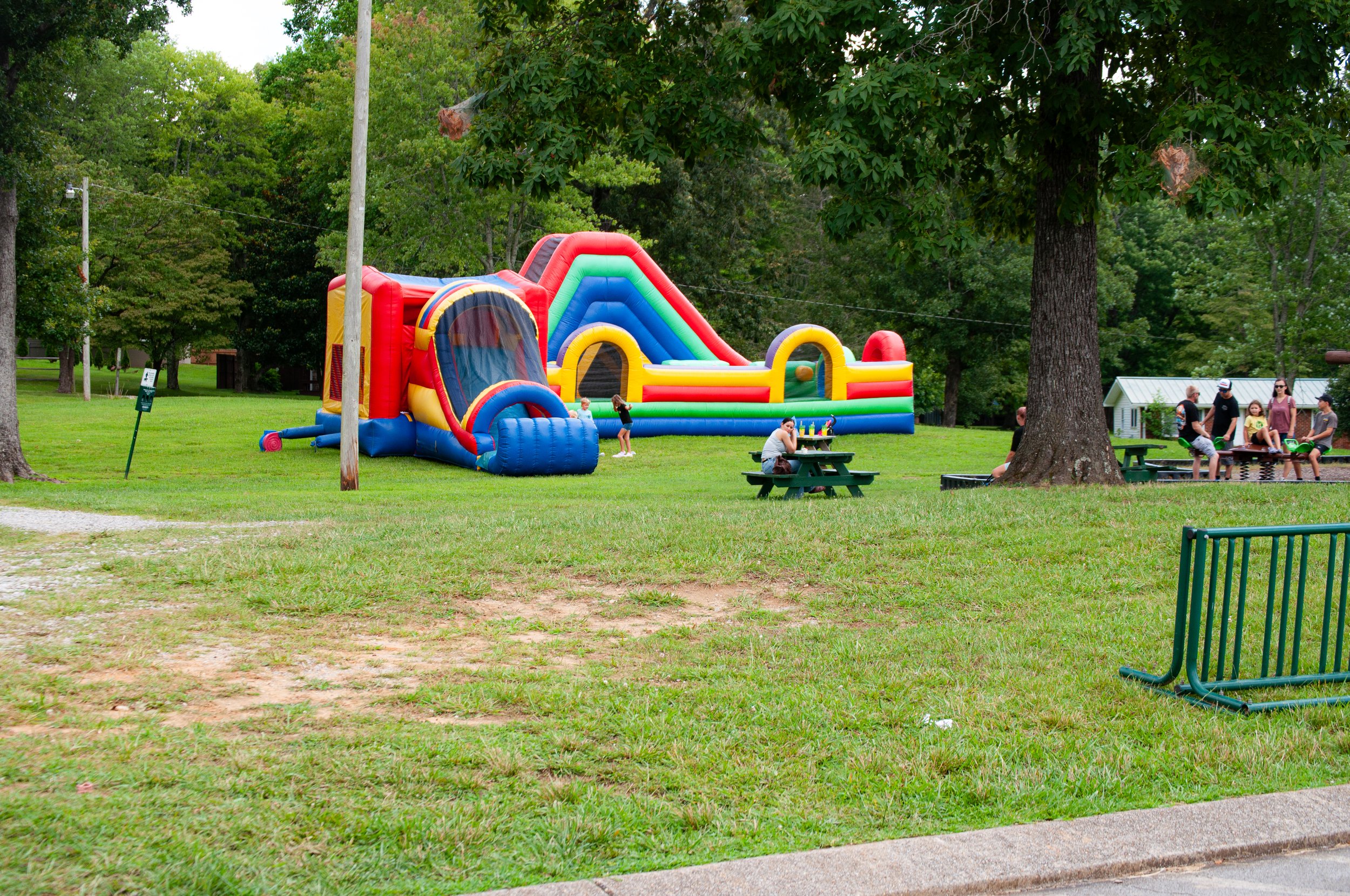 Colorful inflatable bounce house with a slide in a park surrounded by trees, with children and adults nearby