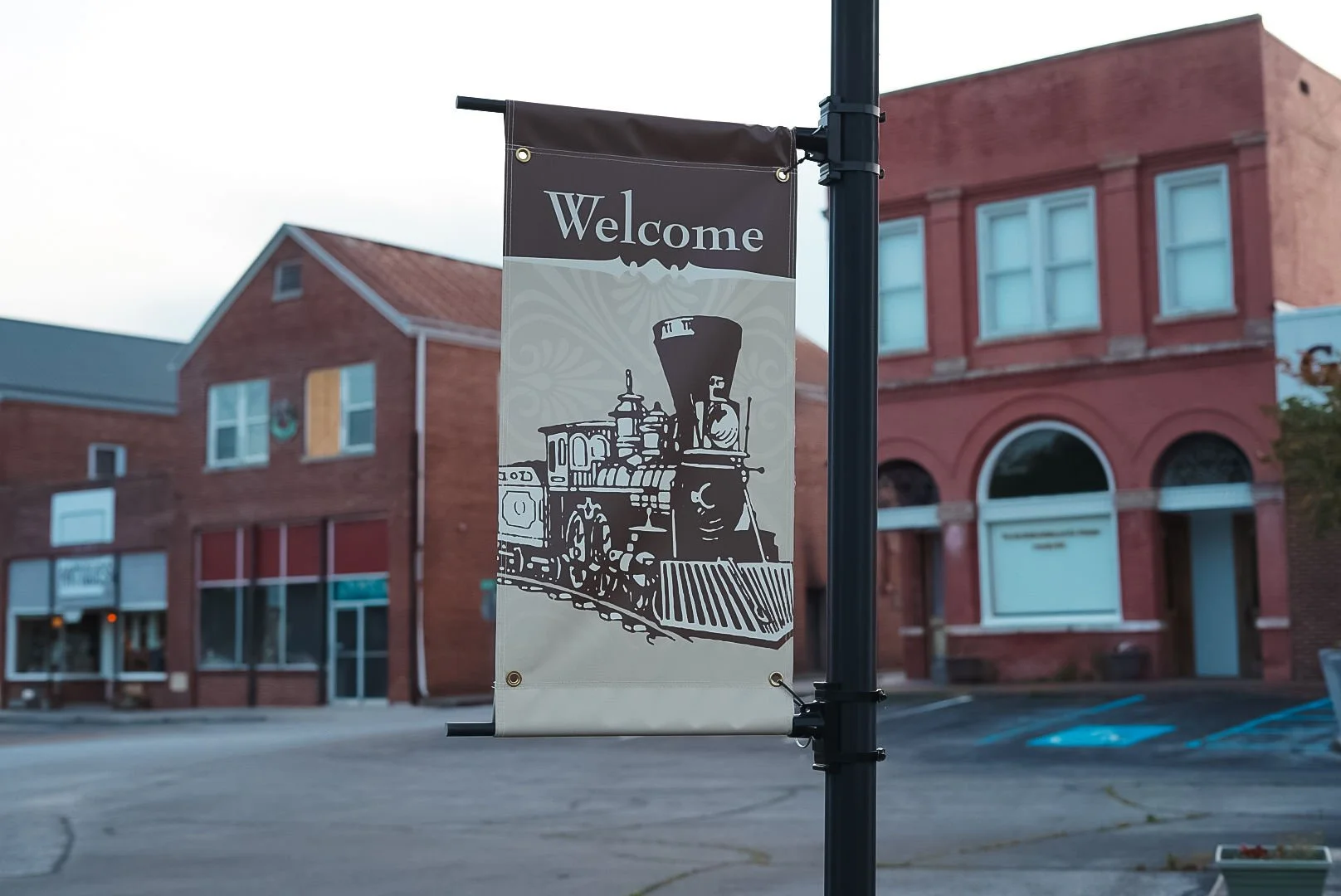Street view with a welcome banner showing an illustration of a steam locomotive train, hanging from a pole in front of brick buildings.