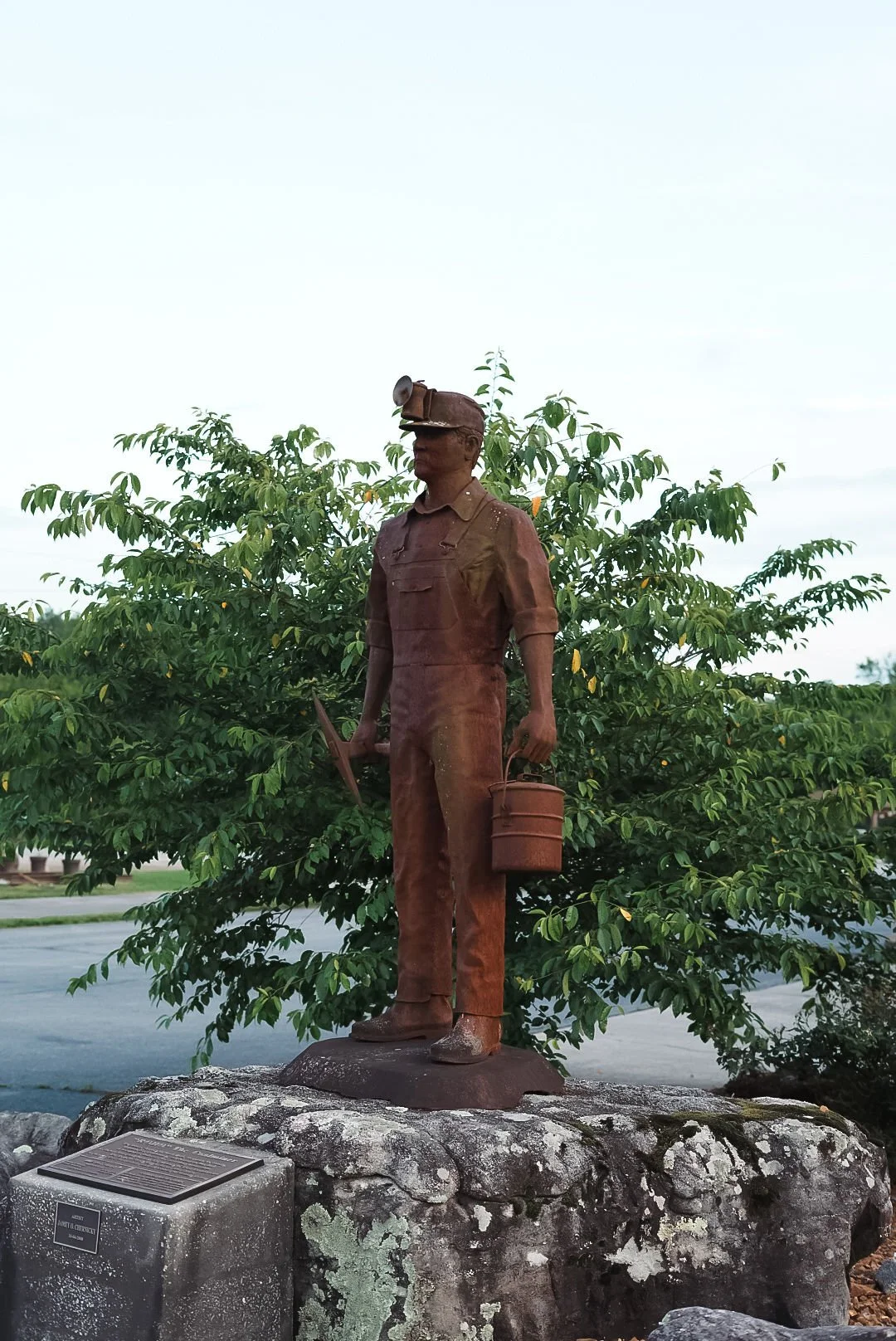 Bronze statue of a man in work clothes holding a bucket and a tool, standing on a rock pedestal with a plaque, outdoors with green foliage in the background.