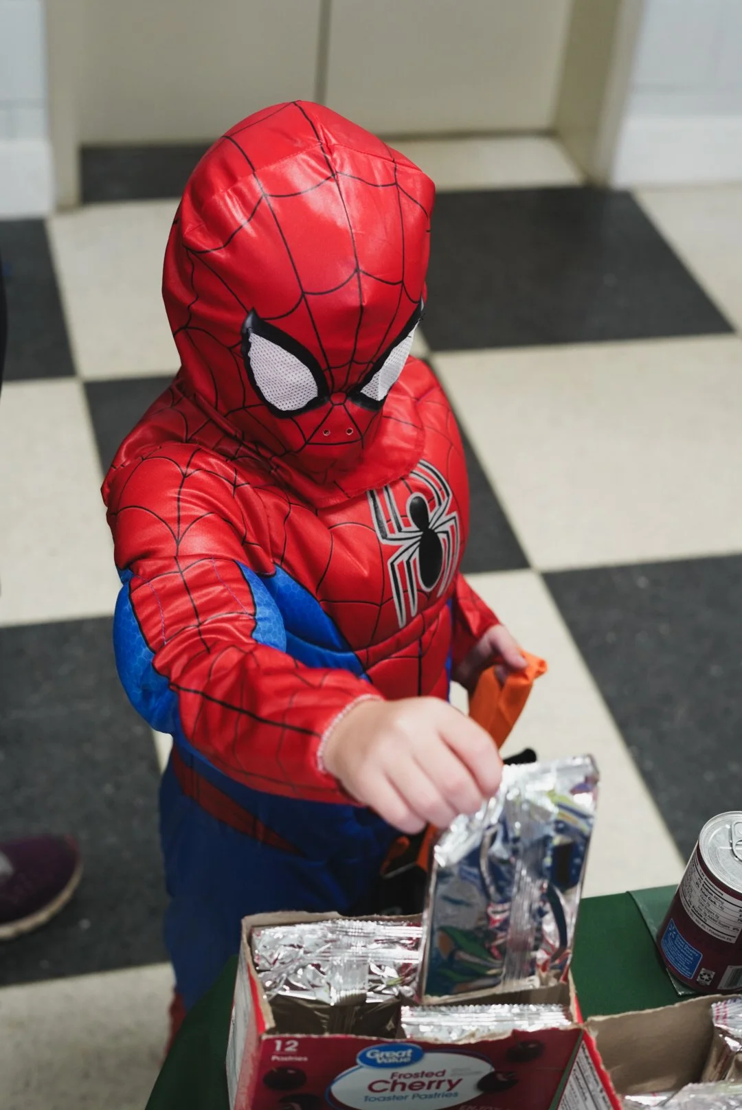 A young child dressed in a Spider-Man costume with a mask, reaching into a box of packaged snacks on a table in a room with black and white checkered flooring.