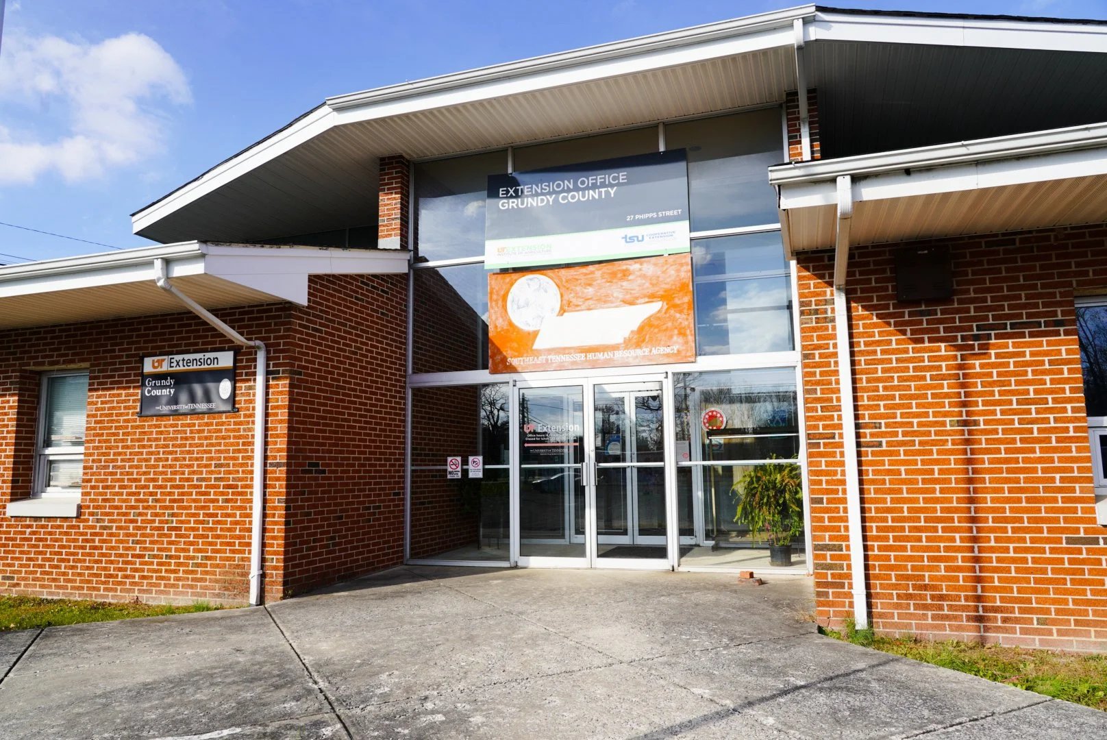 Brick building with glass entrance, signs for Extension Office of Grundy County and Southeast Tennessee Human Resource Agency, and a potted plant inside.
