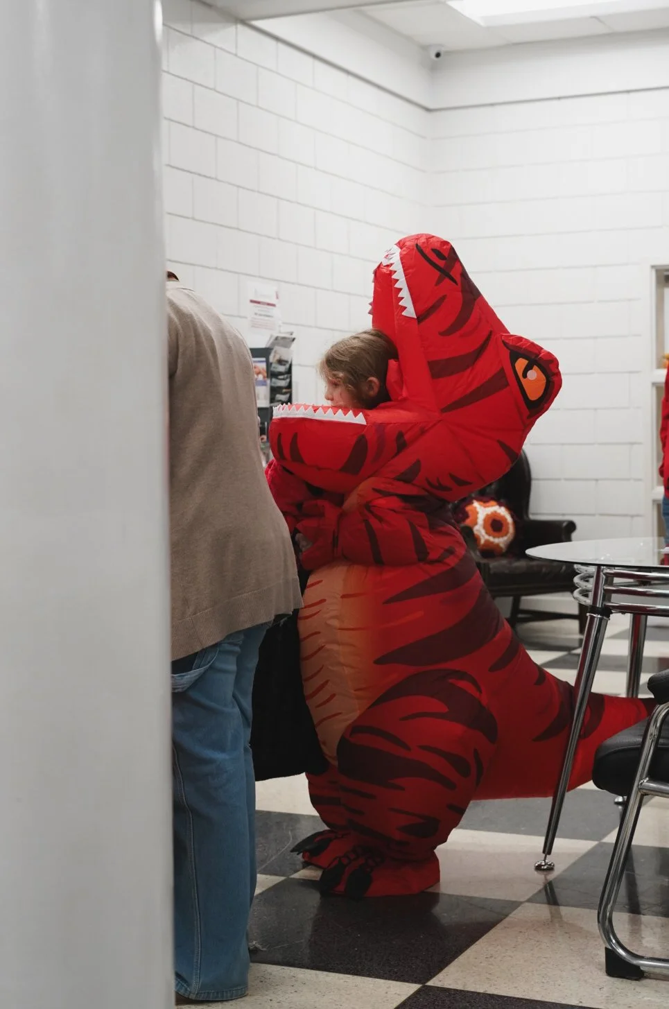 Child in a red tiger costume with a hood, standing in a room with white brick walls, near several people and a black bench with a floral pillow.