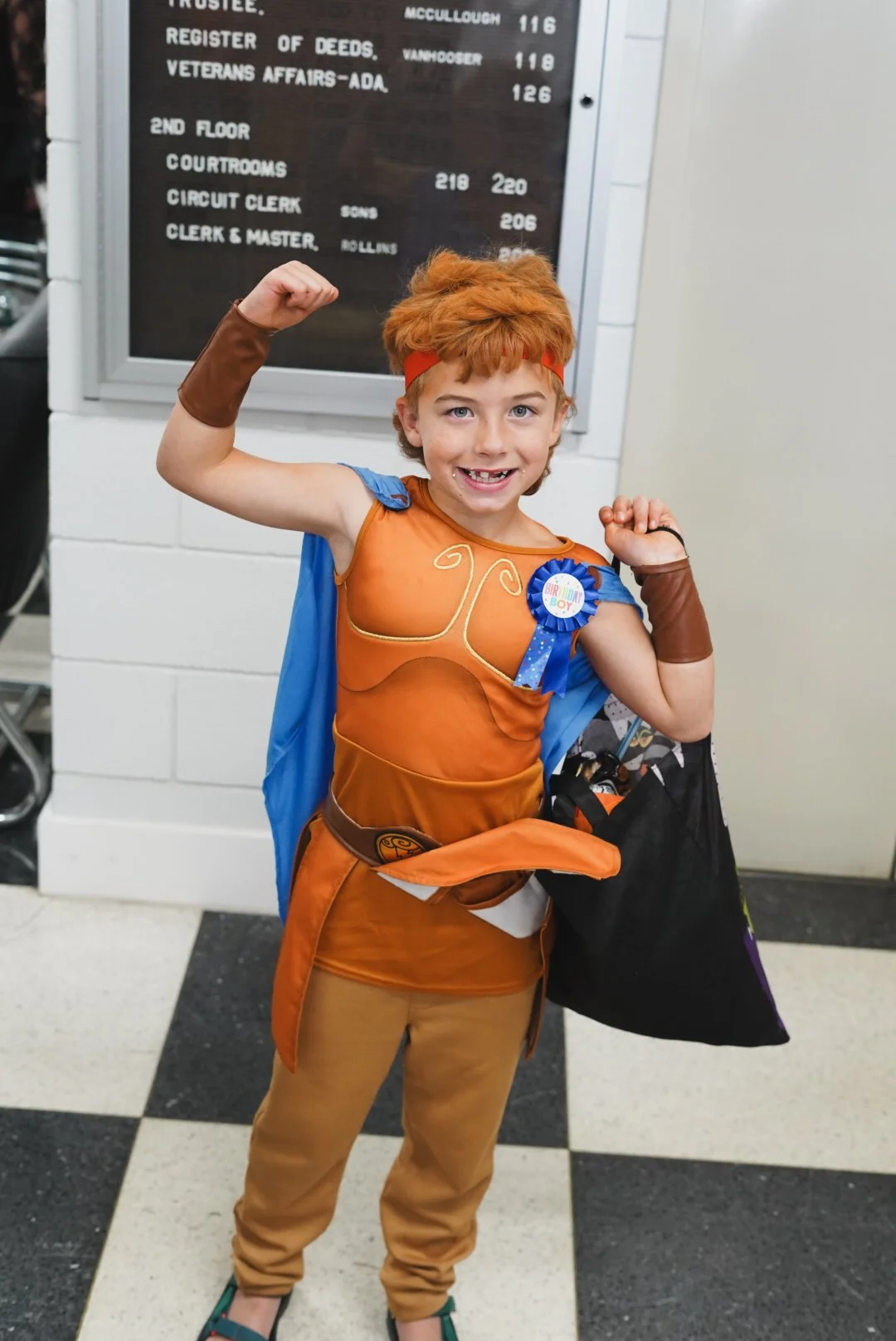 Young boy dressed in a costume, flexing his biceps and smiling at the camera, standing in front of a directory board.