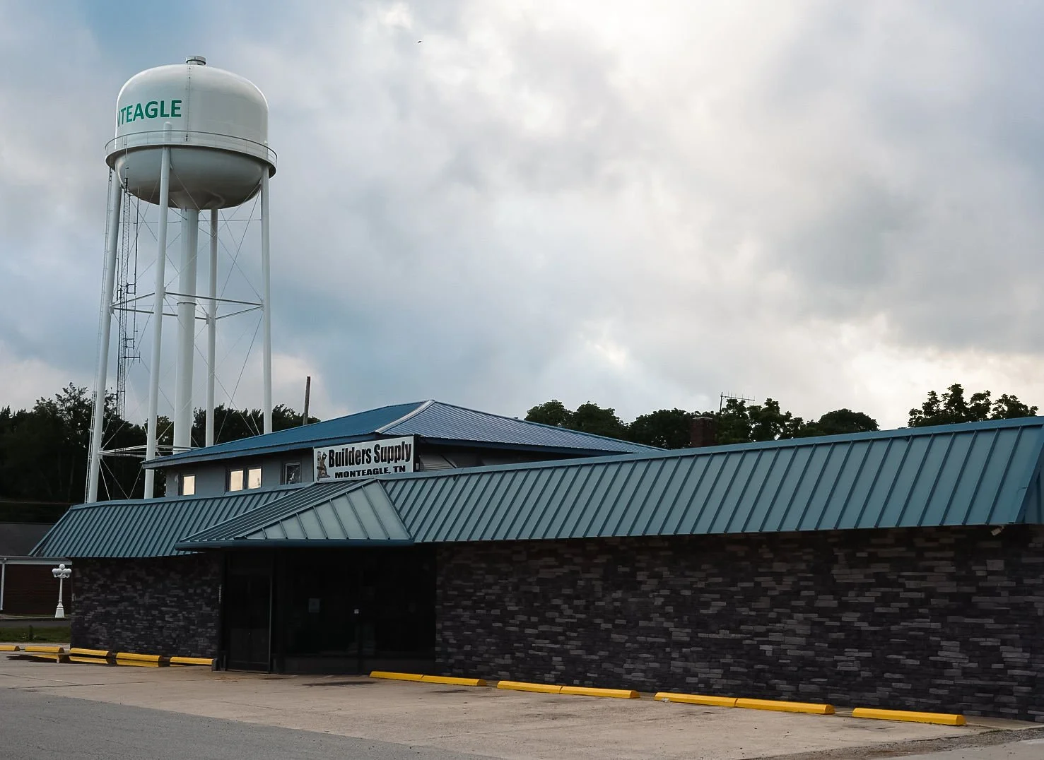 A building with a sign that reads 'Builders Supply' in Monteagle, Tennessee, with a water tower in the background under a cloudy sky.