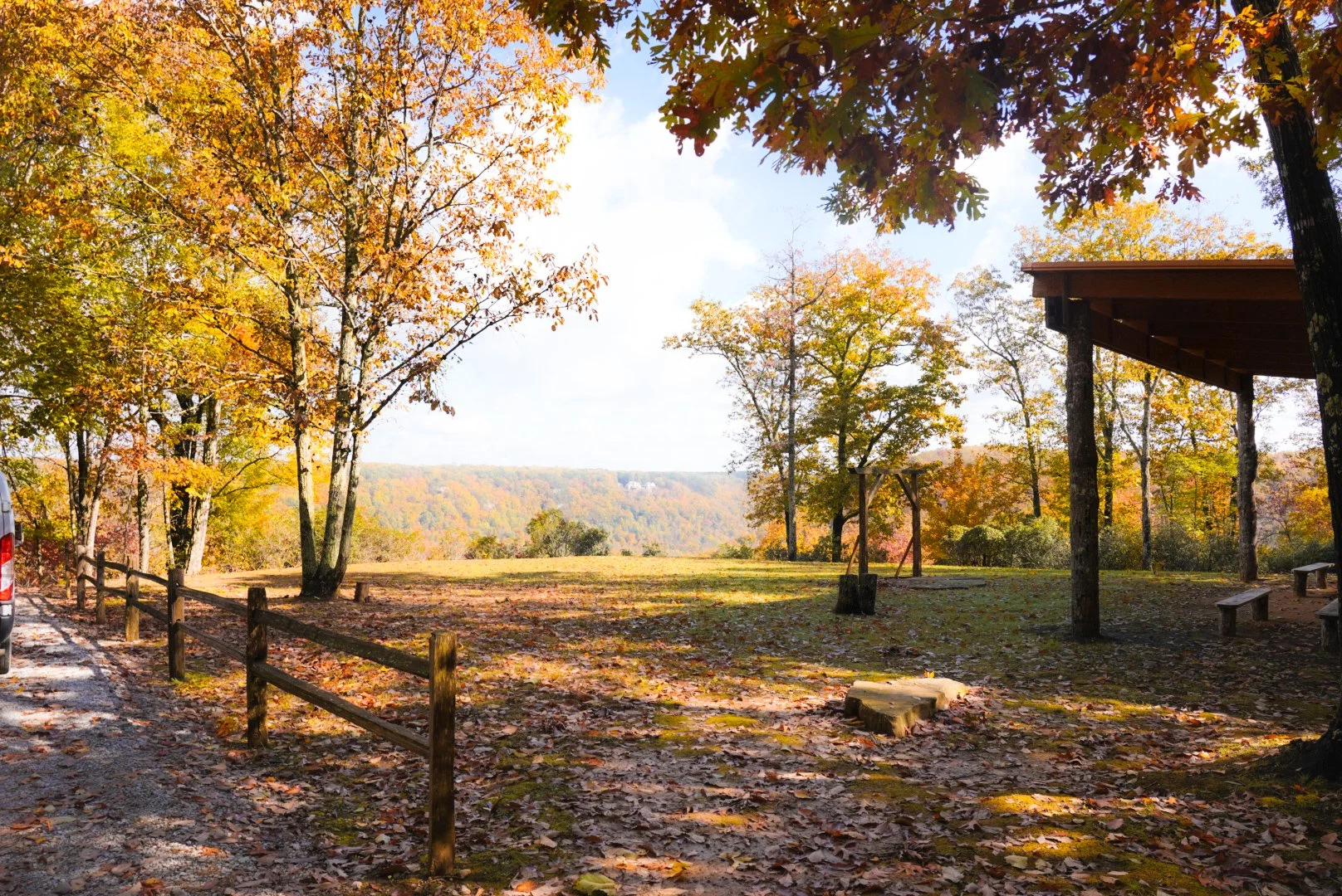 A scenic view of a park or picnic area during autumn with colorful trees, a wooden shelter, and a gravel path. The background features a distant landscape with rolling hills and a clear sky.