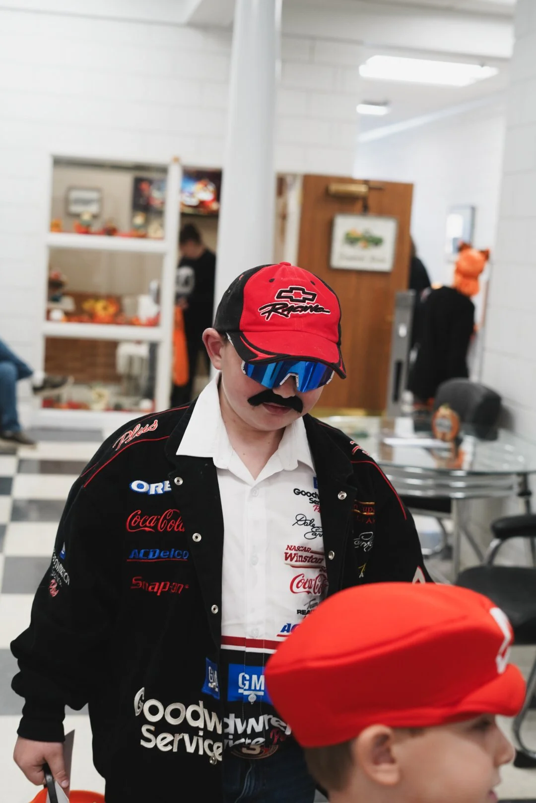 A person dressed in a racing-themed costume, including a red cap, black sunglasses, a fake mustache, a black racing jacket with sponsor logos, and a white shirt underneath, standing indoors with halloween decorations in the background.
