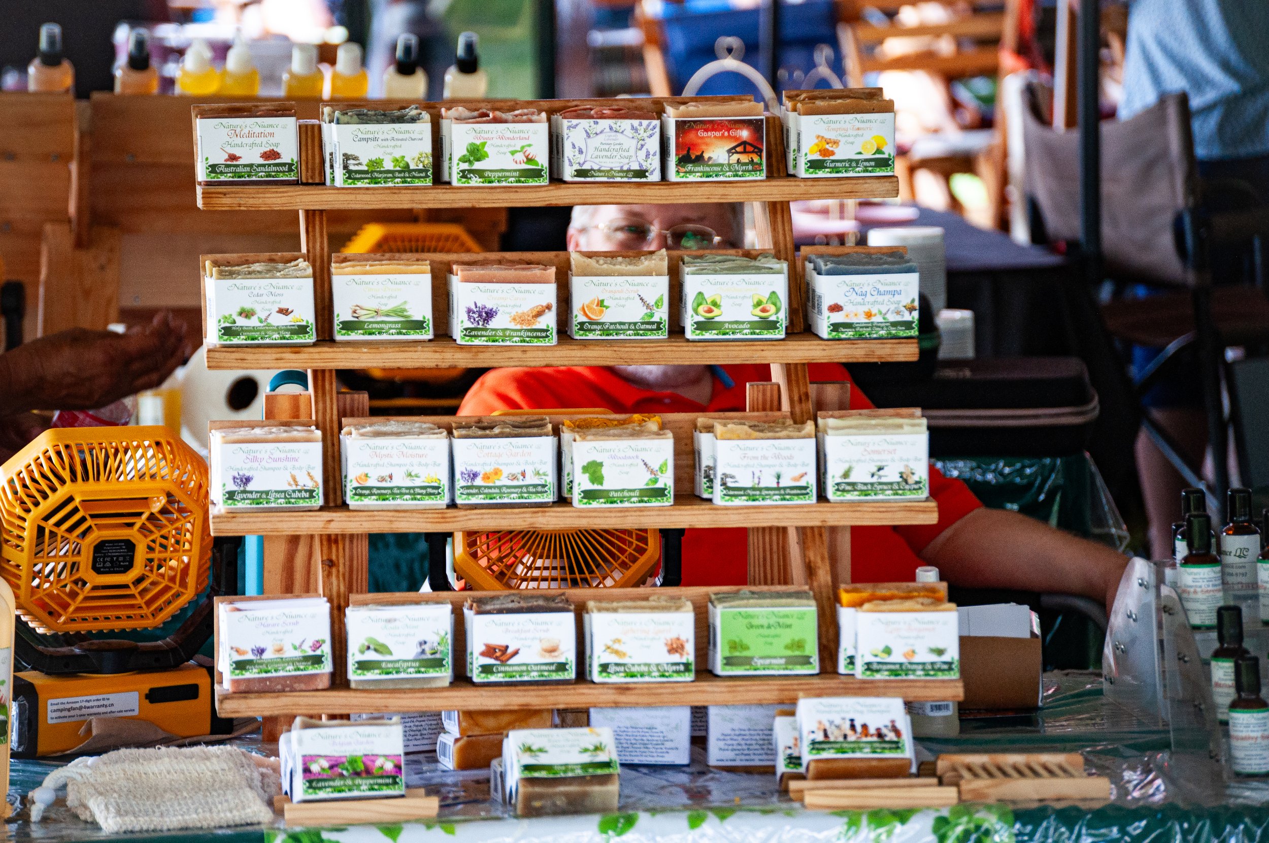 Display of handcrafted soap bars arranged on wooden shelves at an outdoor market, with colorful bottles in the background.