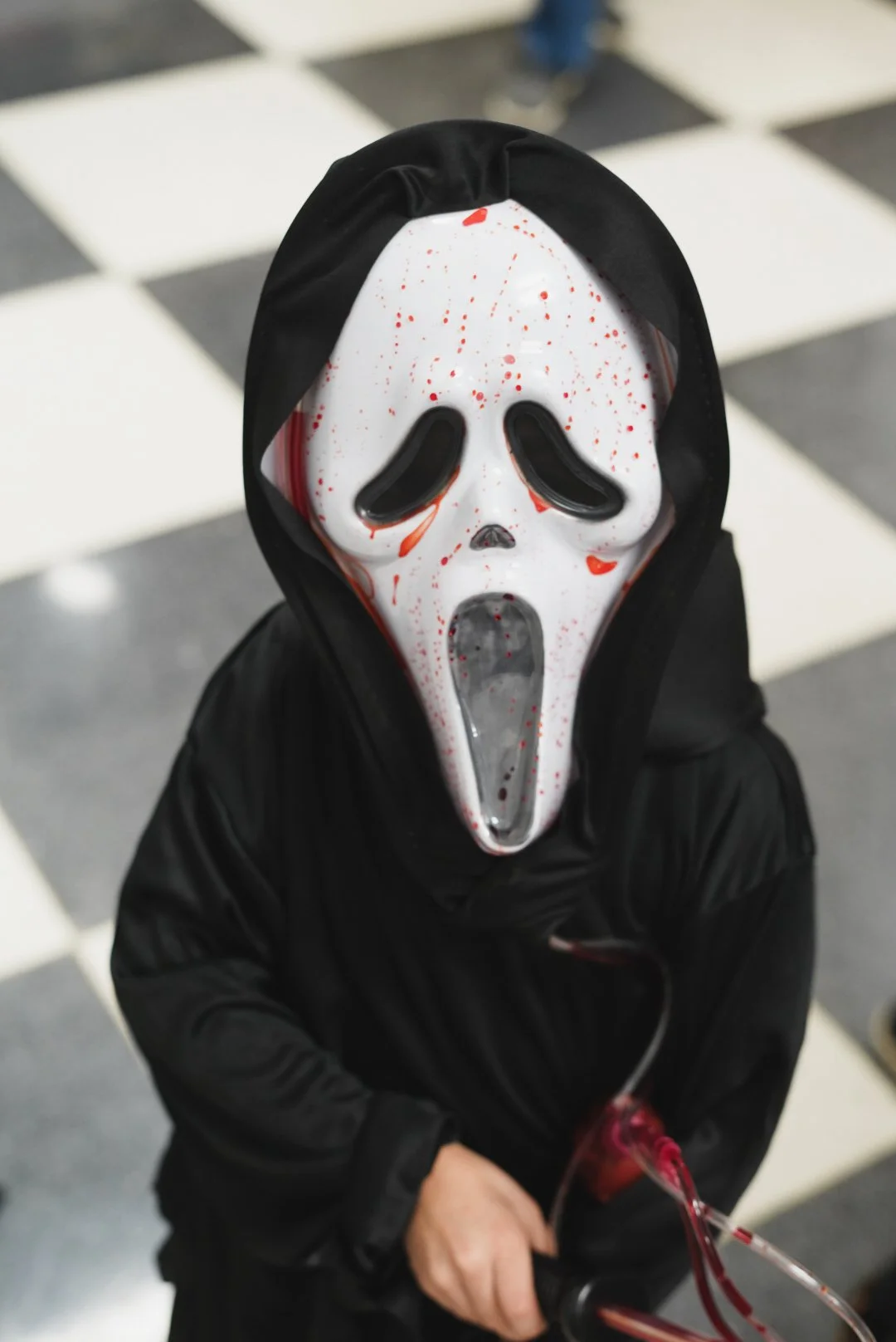 Person wearing a horror-themed mask, black hooded jacket, standing on a checkered floor.